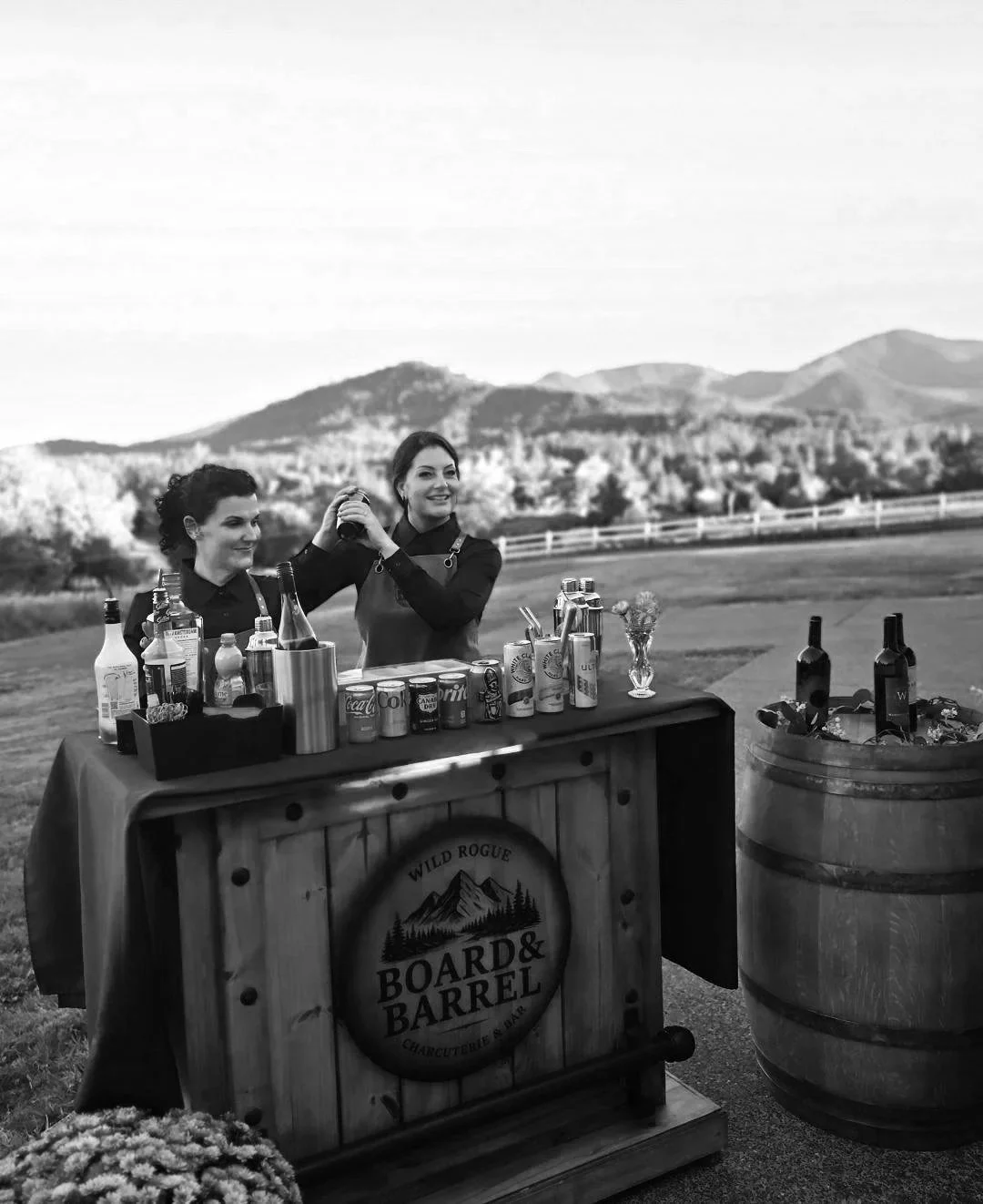 Two women preparing cocktails at an outdoor bar setup with mountains in the background, bottles and cans on the counter, and a wooden sign reading 'Board & Barrel'.