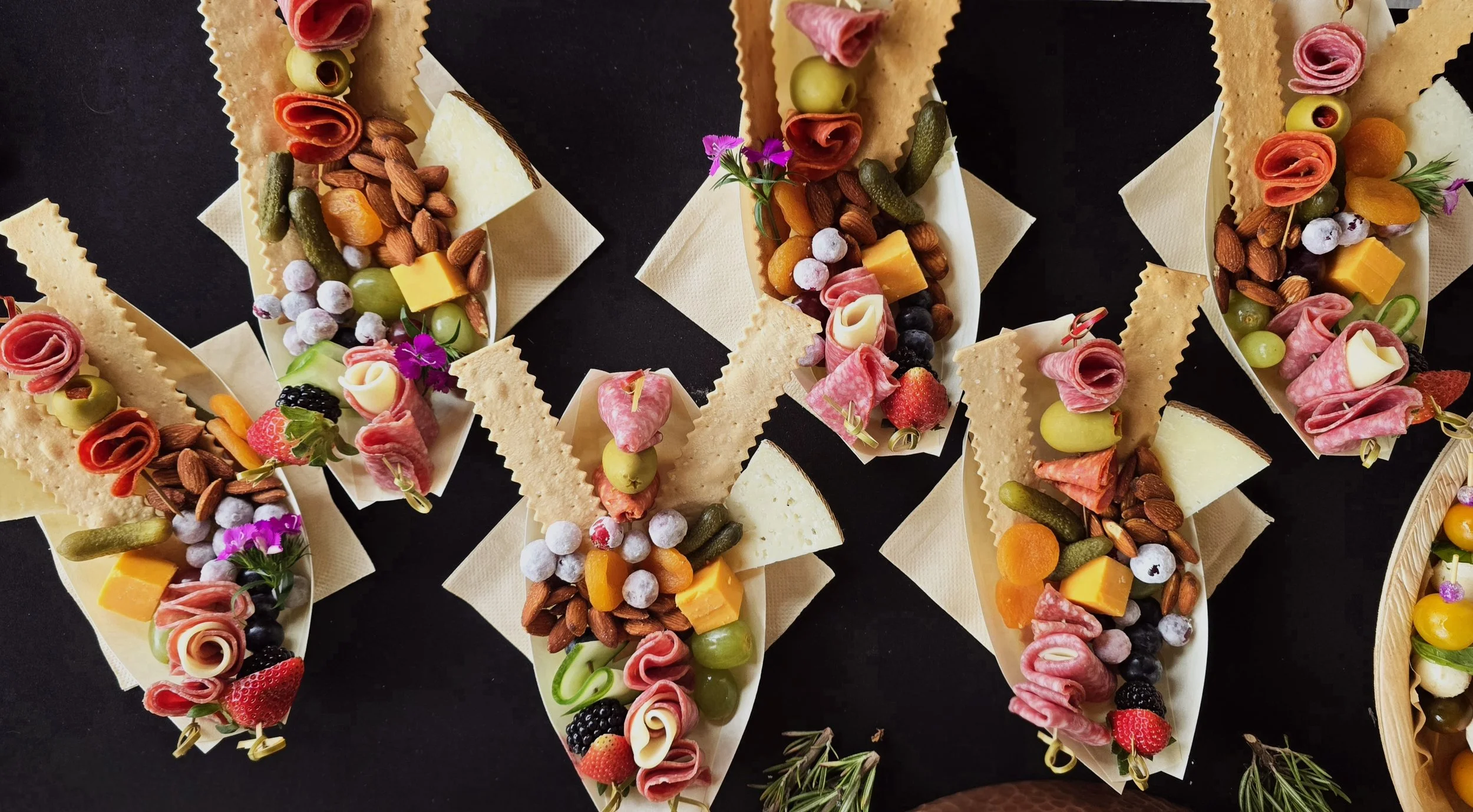 Assorted charcuterie boats with cheeses, meats, fruits, nuts, and garnishes on a black background.