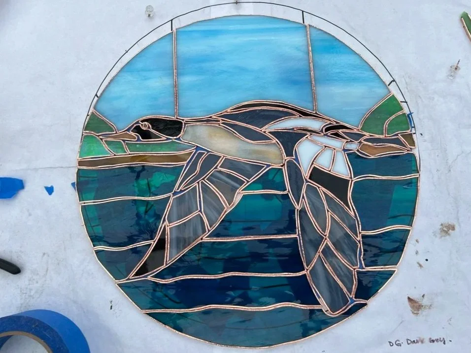 A stained glass window depicting a bird standing in water with green land and blue sky in the background.