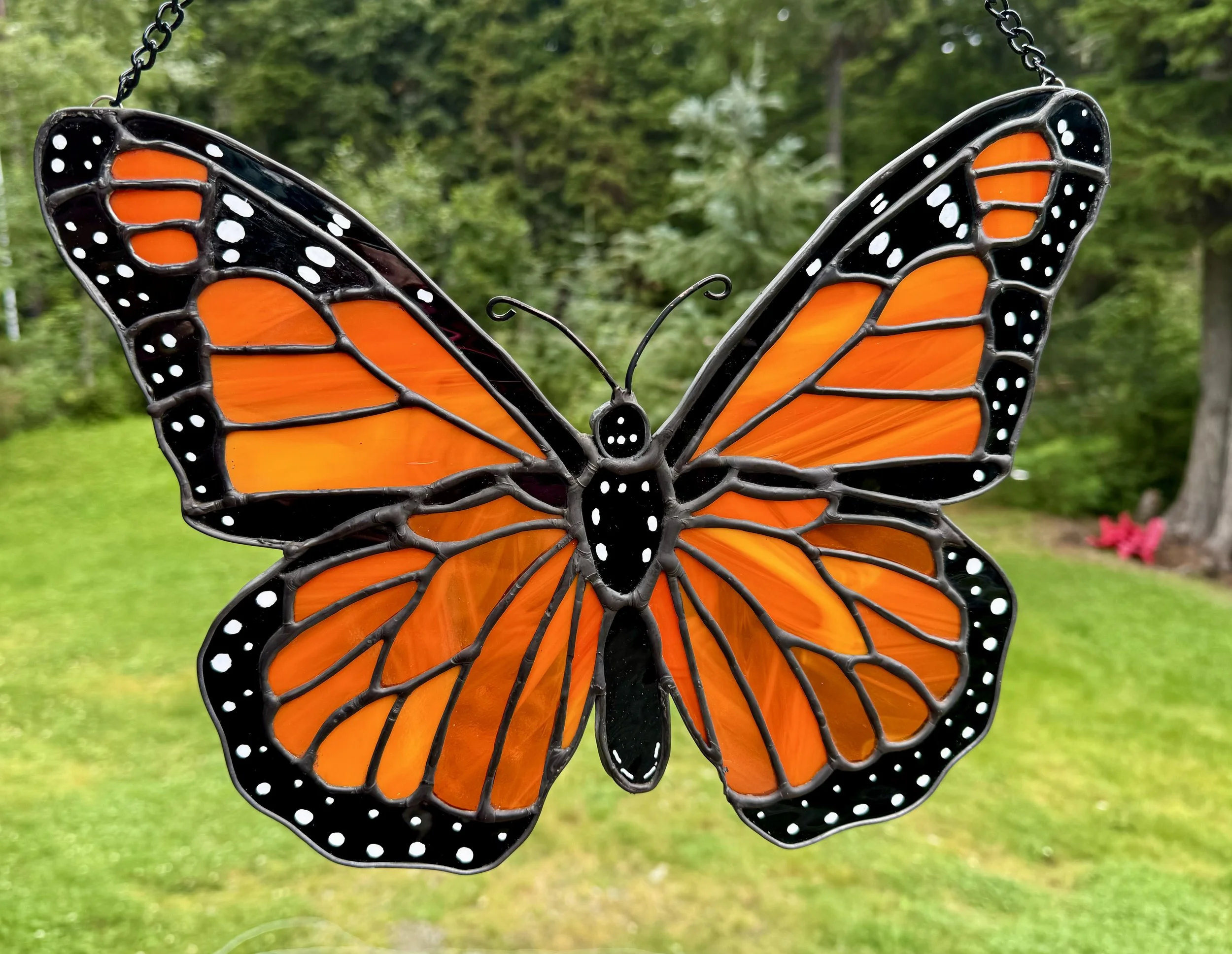 A stained glass butterfly with orange and black wings, suspended outdoors against a green background.