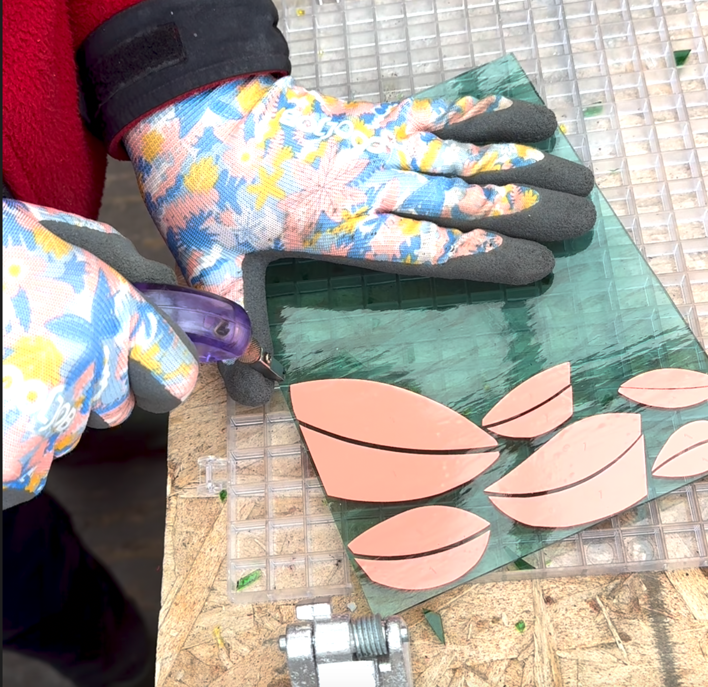 A person wearing floral-patterned gloves working on a craft project involving pink vinyl and a green surface with leaf shapes on a wooden table.