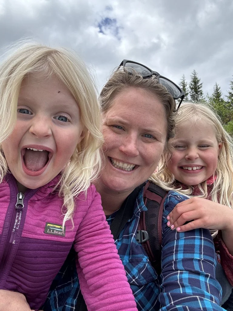 A smiling woman with two young girls, all with blonde hair, outdoors with trees and cloudy sky in the background.