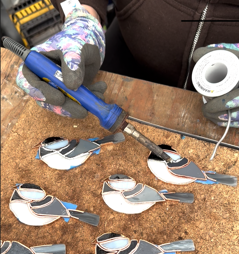 A person wearing gloves and a brown jacket is soldering bird-shaped ornaments on a wooden surface. The person is holding a soldering iron, and a spool of solder wire is nearby.