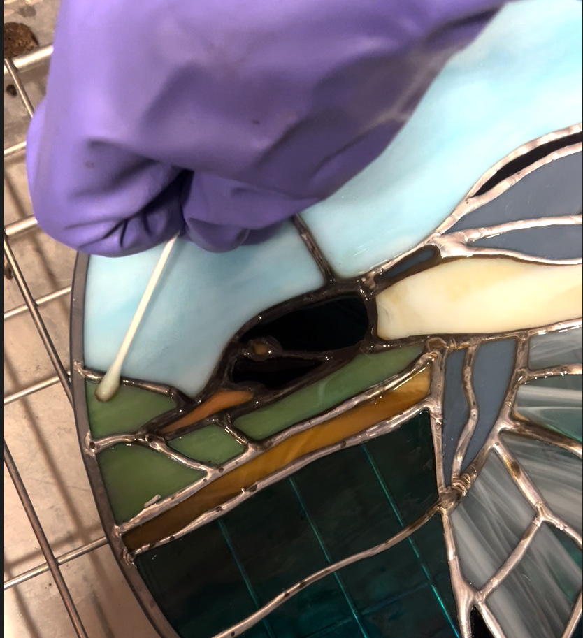 Close-up of a stained glass art piece depicting an eye being assembled, with a person's gloved hand holding a cotton swab nearby.
