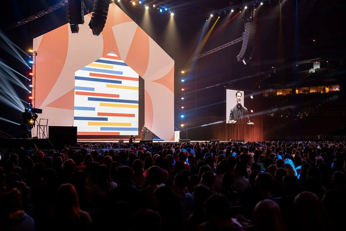 A speaker on stage at a conference or event, with a large digital screen behind them displaying a colorful abstract design, and a smaller screen showing the speaker's face, surrounded by an audience in a darkened auditorium.