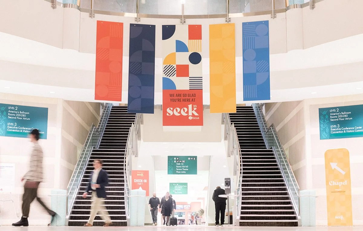 Interior view of a conference center with two staircases, colorful banners hanging from the ceiling, and people walking around. Signs indicate levels, conference rooms, and check-in areas.