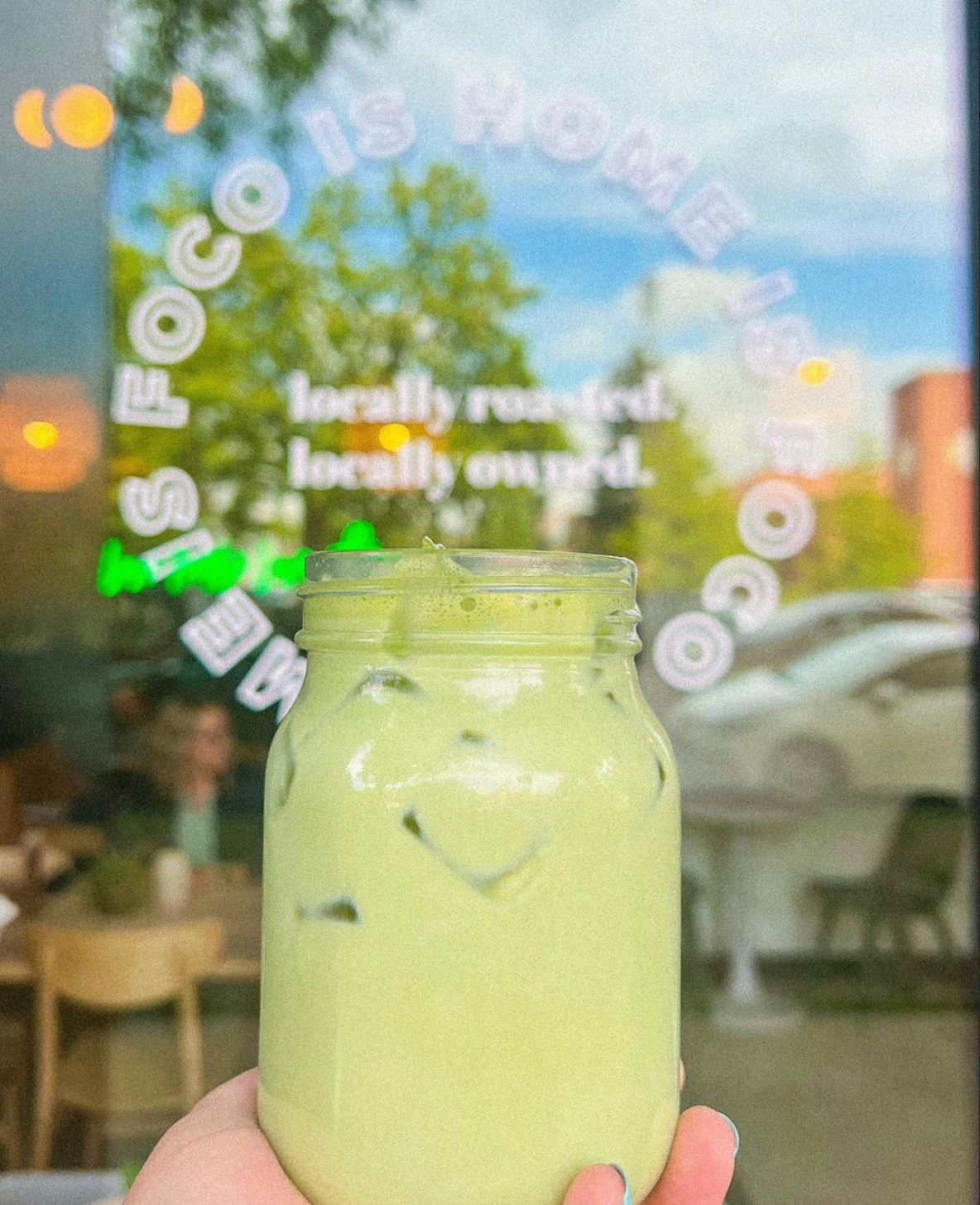 A person holding a mason jar filled with a light green beverage with ice, in front of a glass window with the words 'COFFEE SHOP' and menu items written on it, with outdoor seating and people inside the cafe visible through the window.