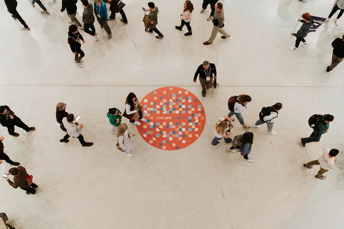 People walking through a large indoor space with one circle on the floor that says 'you are called'.