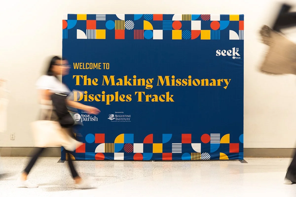 A large blue poster with colorful geometric shapes at the top and bottom, reading 'Welcome to The Making Missionary Disciples Track,' sponsored by Focus Parish and Augustine Institute, displayed in an indoor setting with blurred people walking by.
