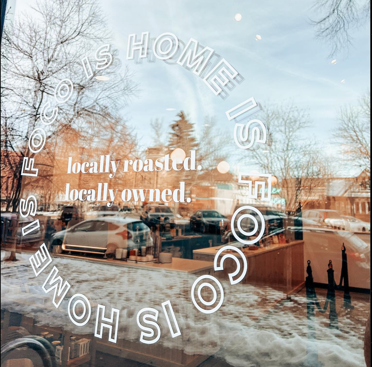 View through a glass window with white text that reads 'FOCO IS HOME IS SPECIAL TO US', 'locally roasted.', 'locally owned.' Reflecting a street scene with cars parked outside, leafless trees, buildings, and snow on the ground during winter.