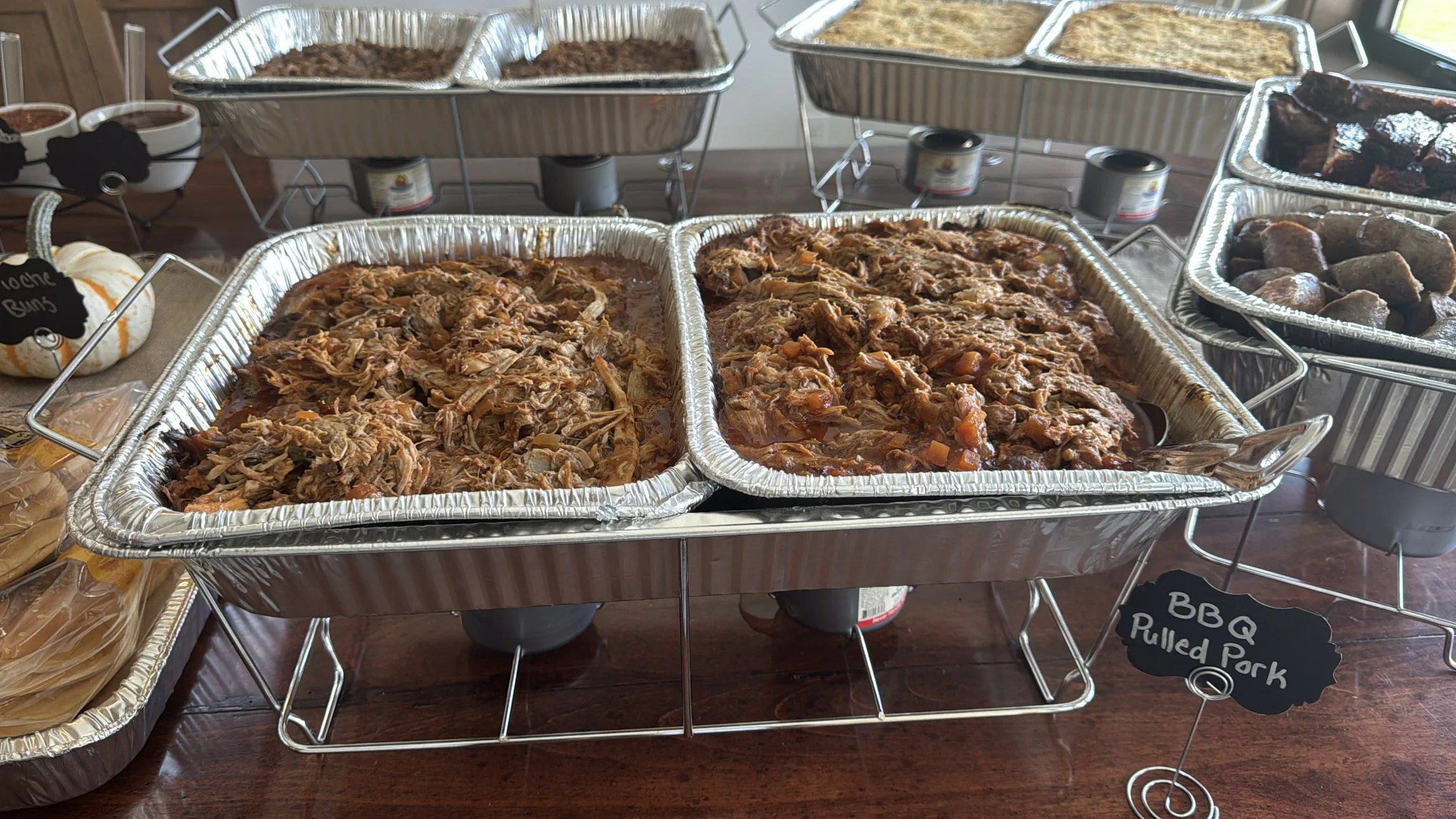 Buffet table with trays of BBQ pulled pork, baked beans, and meatballs, labeled with small chalkboard signs.
