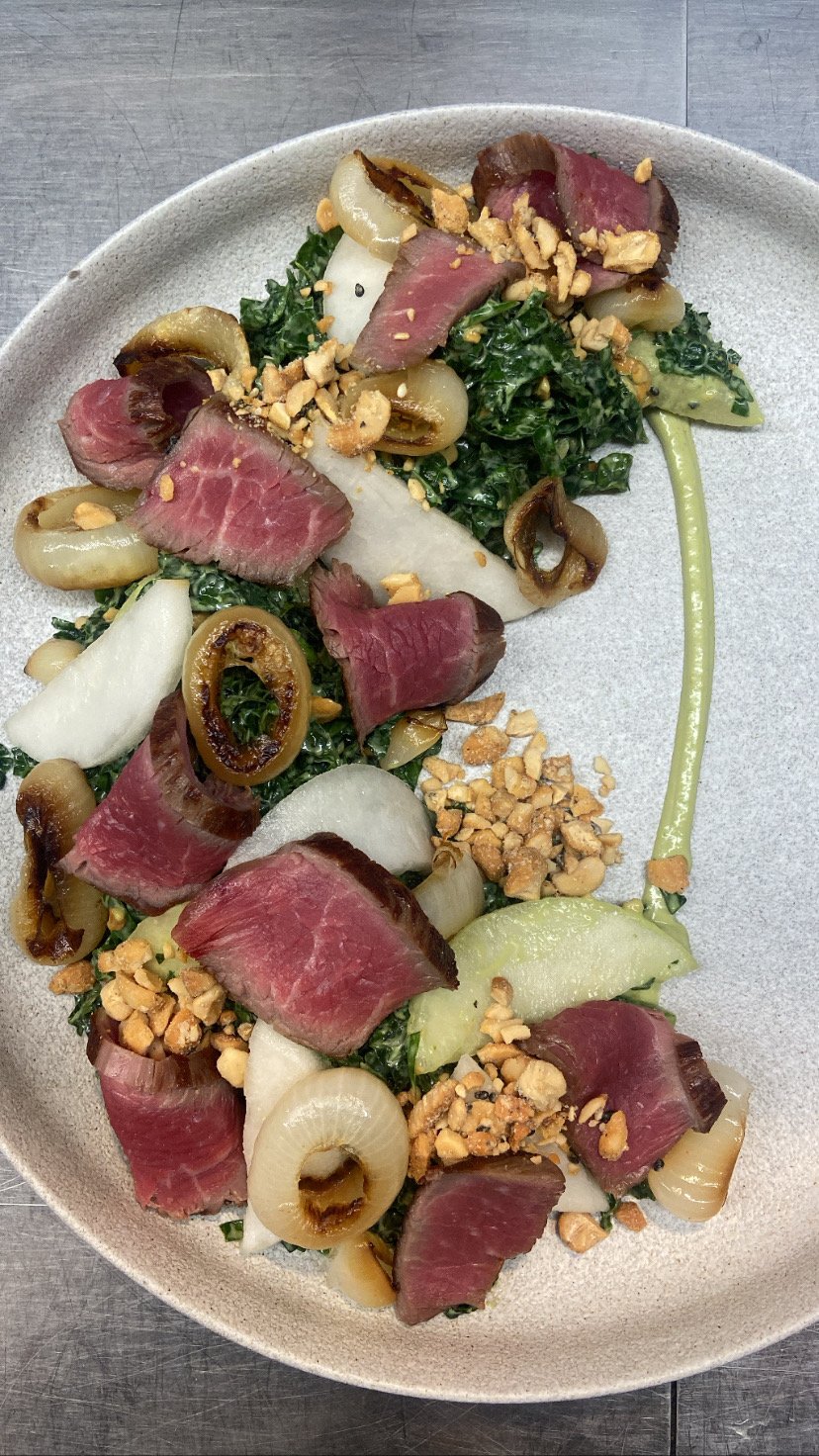 Seared tuna steak with kale, fennel, olives, onions, chopped nuts, and a green sauce on a white oval plate.