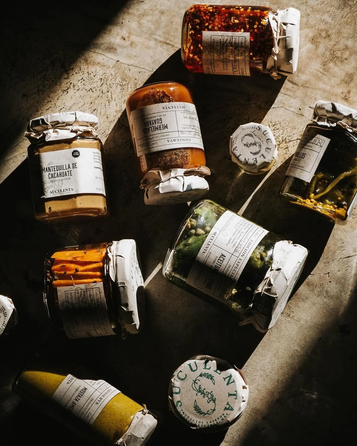 Collection of assorted jars and bottles with labels, containing various preserved foods and condiments, arranged on a textured surface with sunlight casting shadows.