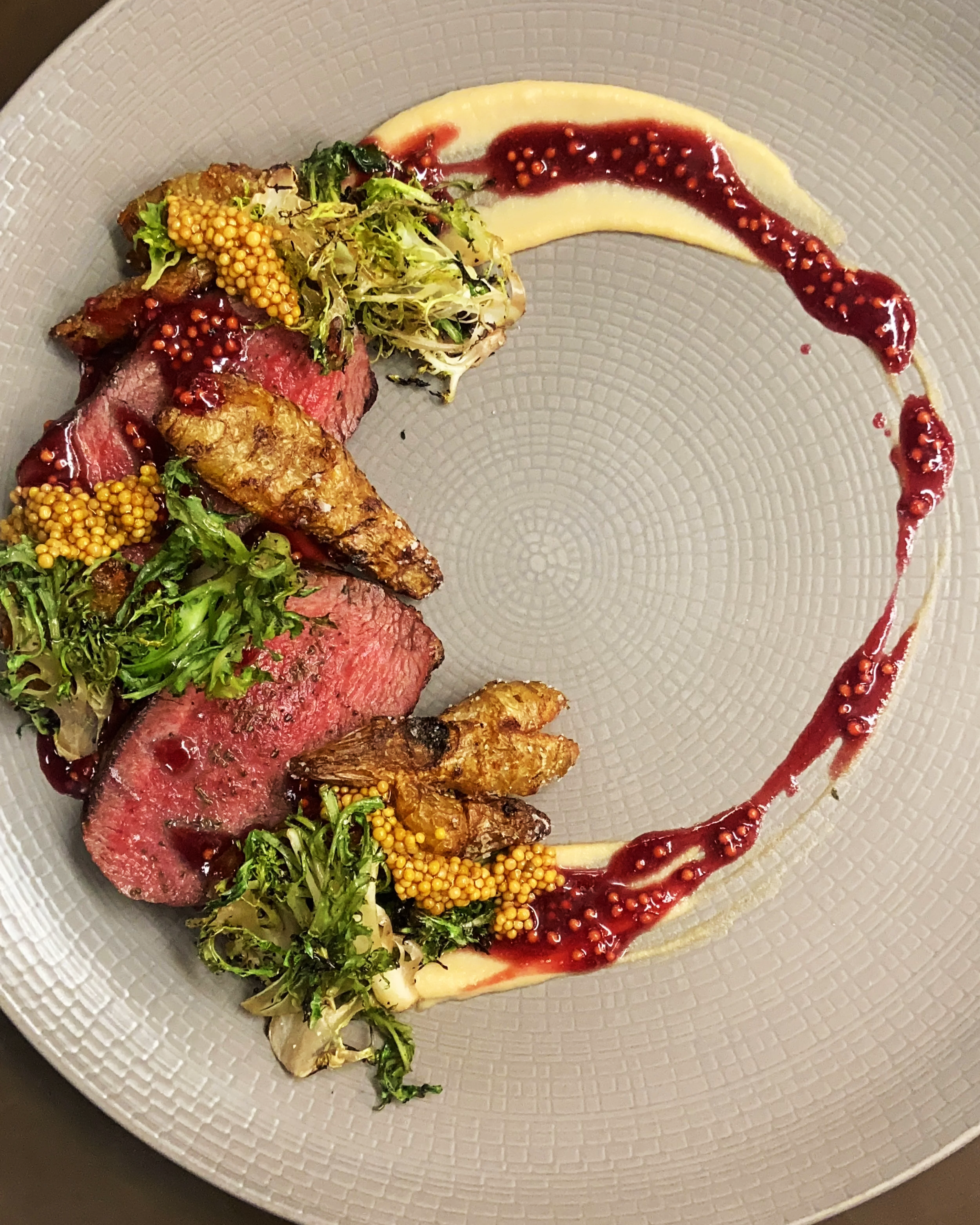 Slices of medium-rare beef steak topped with mustard seeds, arugula, and pickled vegetables, with a smear of white puree and garnished with a red berry sauce on a decorative white plate.