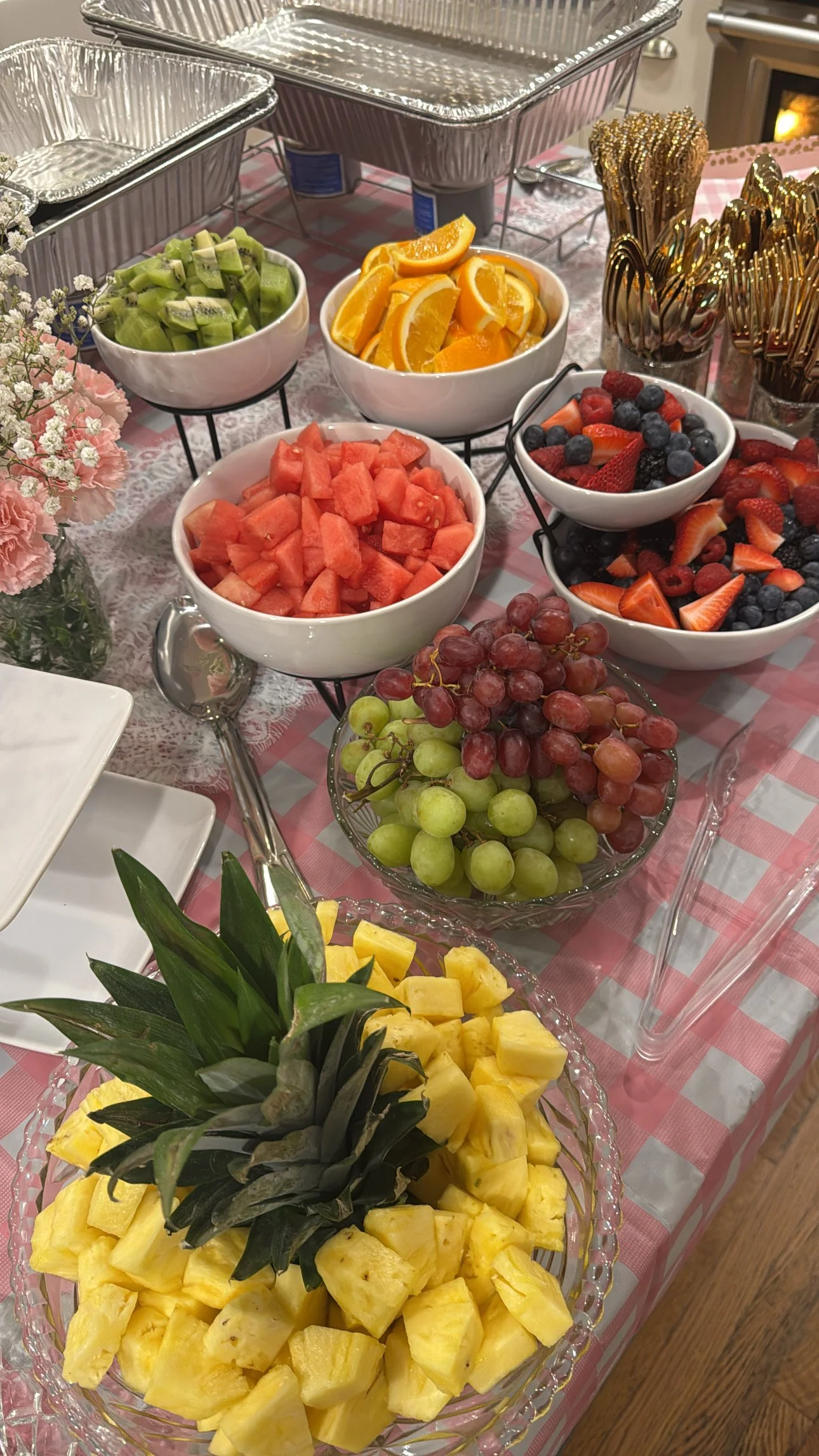 A table of fresh fruit including pineapple, grapes, watermelon, strawberries, blueberries, raspberries, oranges, and kiwi, with serving utensils and floral decorations.