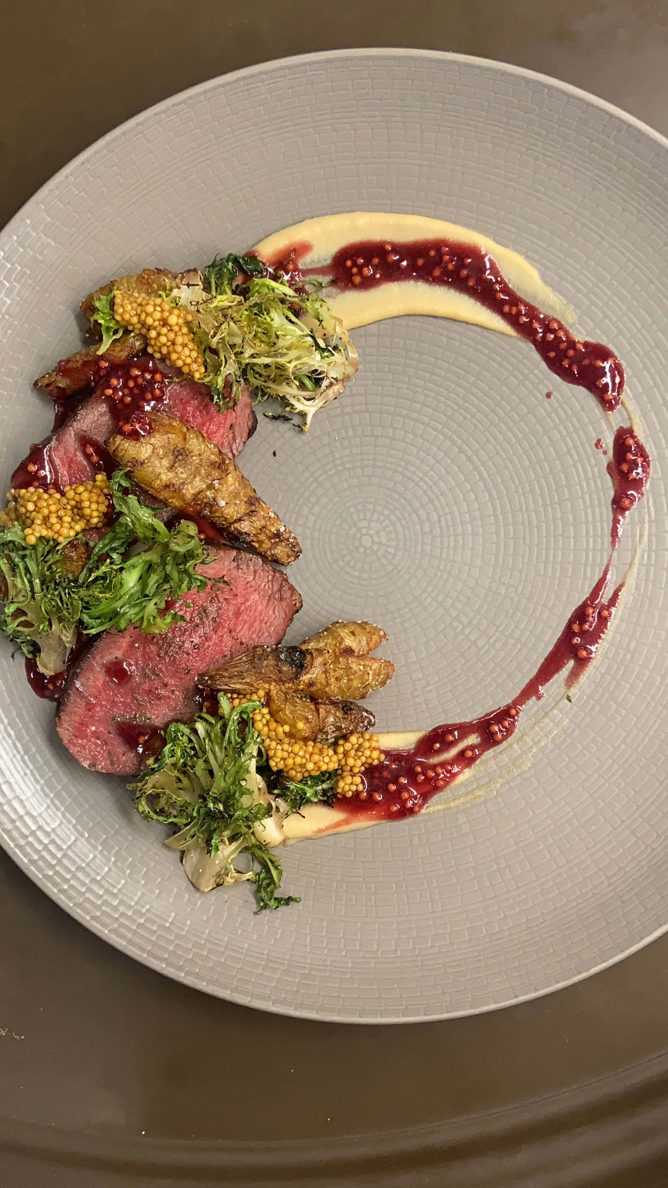 Sliced medium-rare red meat steak topped with greens, mustard seeds, and crispy fried garnish, served with a mustard and berry sauce on a grey textured plate.