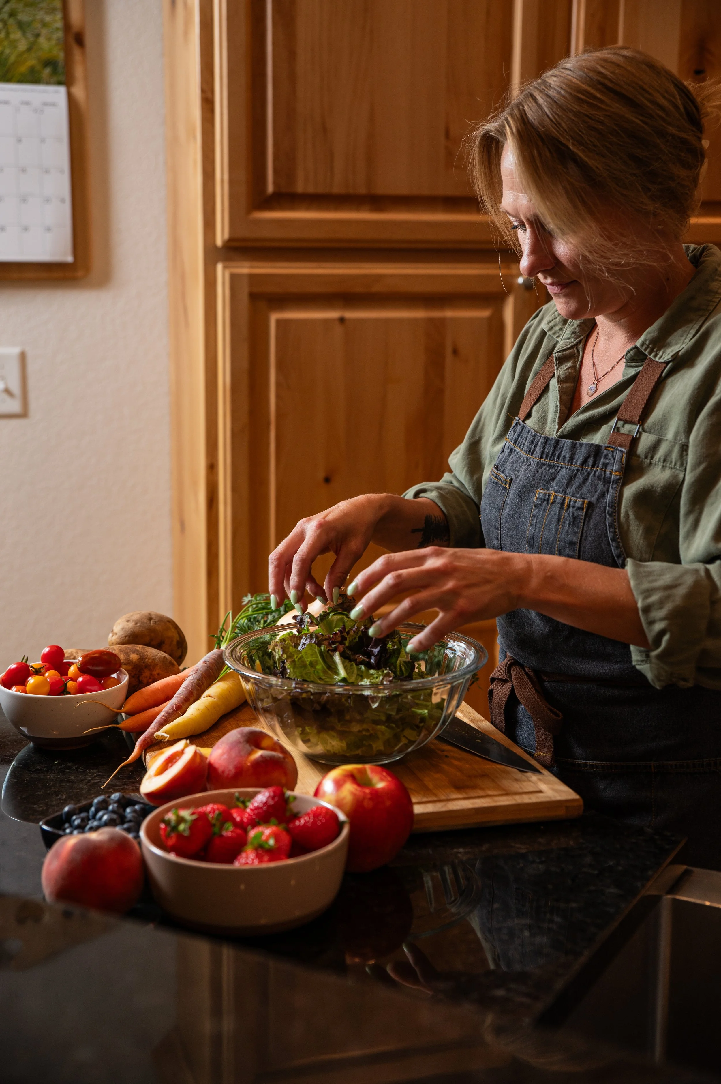A woman preparing a salad in a kitchen with various fresh vegetables and fruits on the counter.