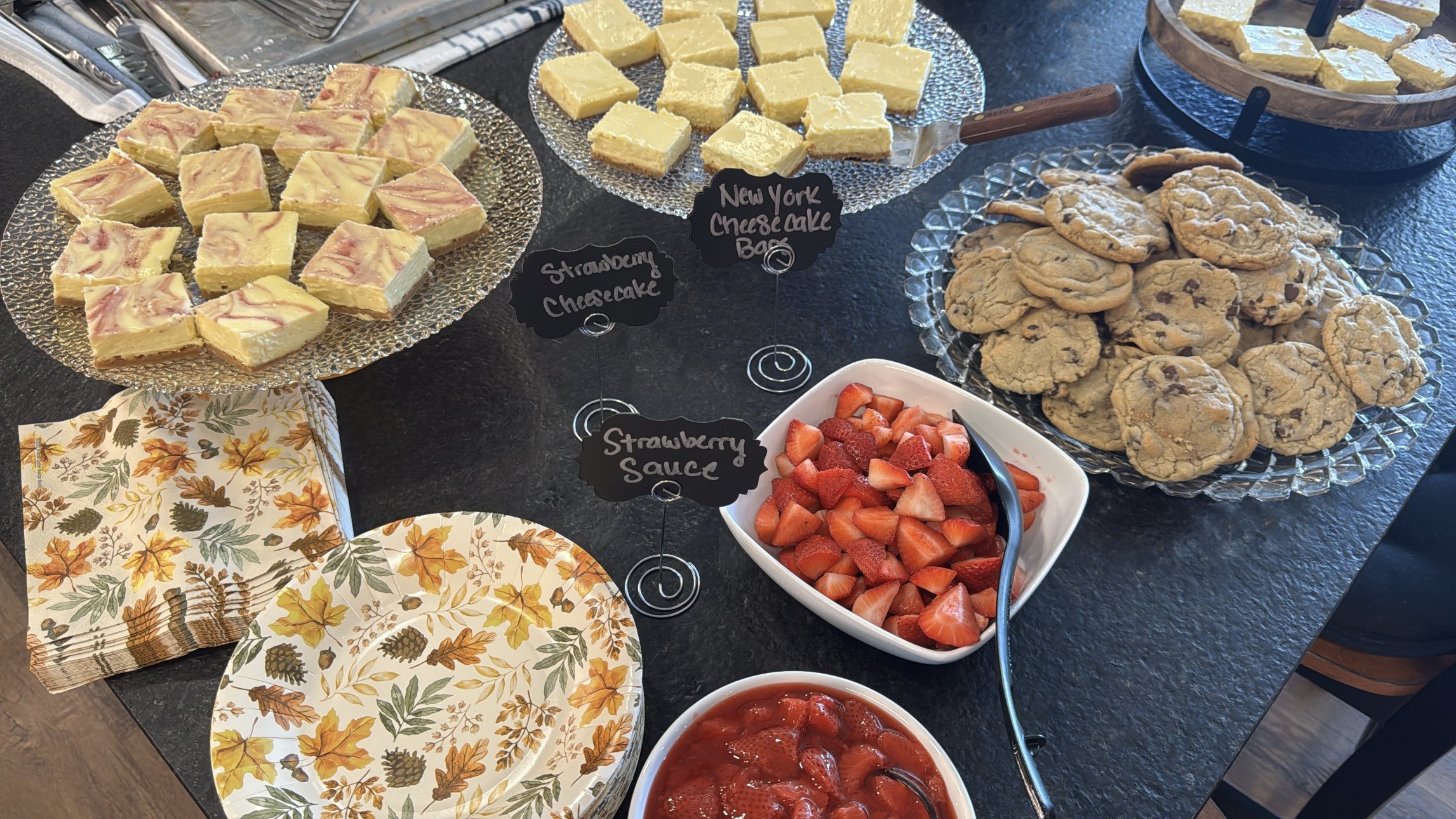 An assortment of desserts and strawberries on a table. There are square slices of strawberry cheesecake, New York cheesecake, chocolate chip cookies, and bowls of sliced strawberries and strawberry sauce, with small black signs labeling each item.