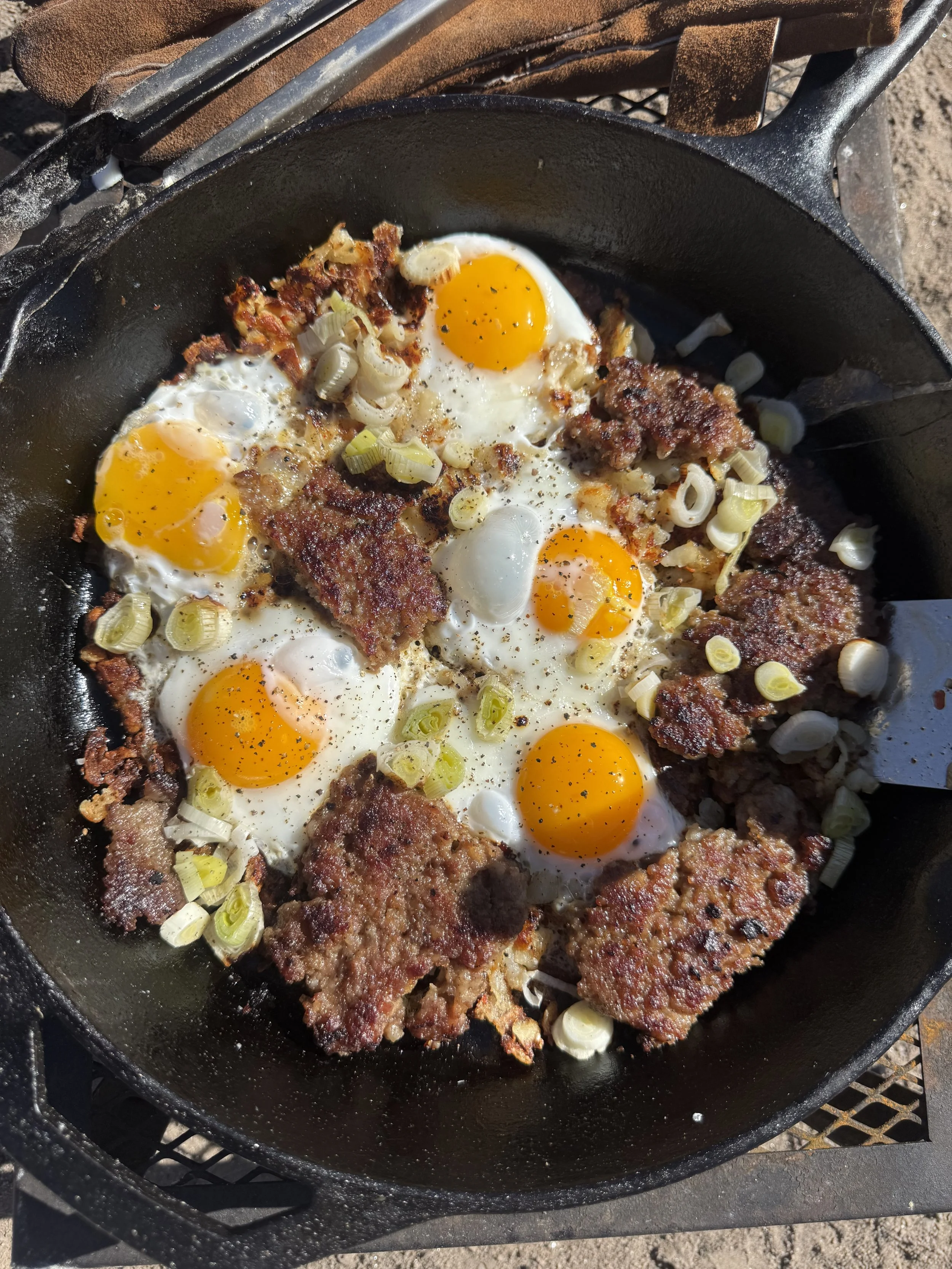 A cast iron skillet with sunny-side-up eggs, cooked sausage, and chopped green onions on a grill outdoors.