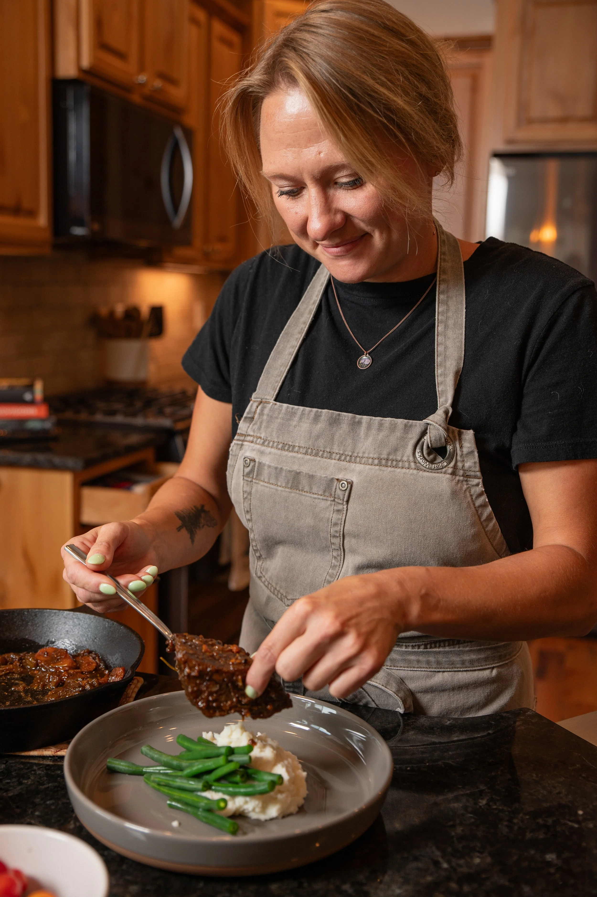A woman with short red hair wearing a black t-shirt, beige apron, and a silver necklace preparing food in a kitchen with wooden cabinets. She is placing a cooked piece of meat or a similar dish onto a plate with green beans, mashed potatoes, and a do