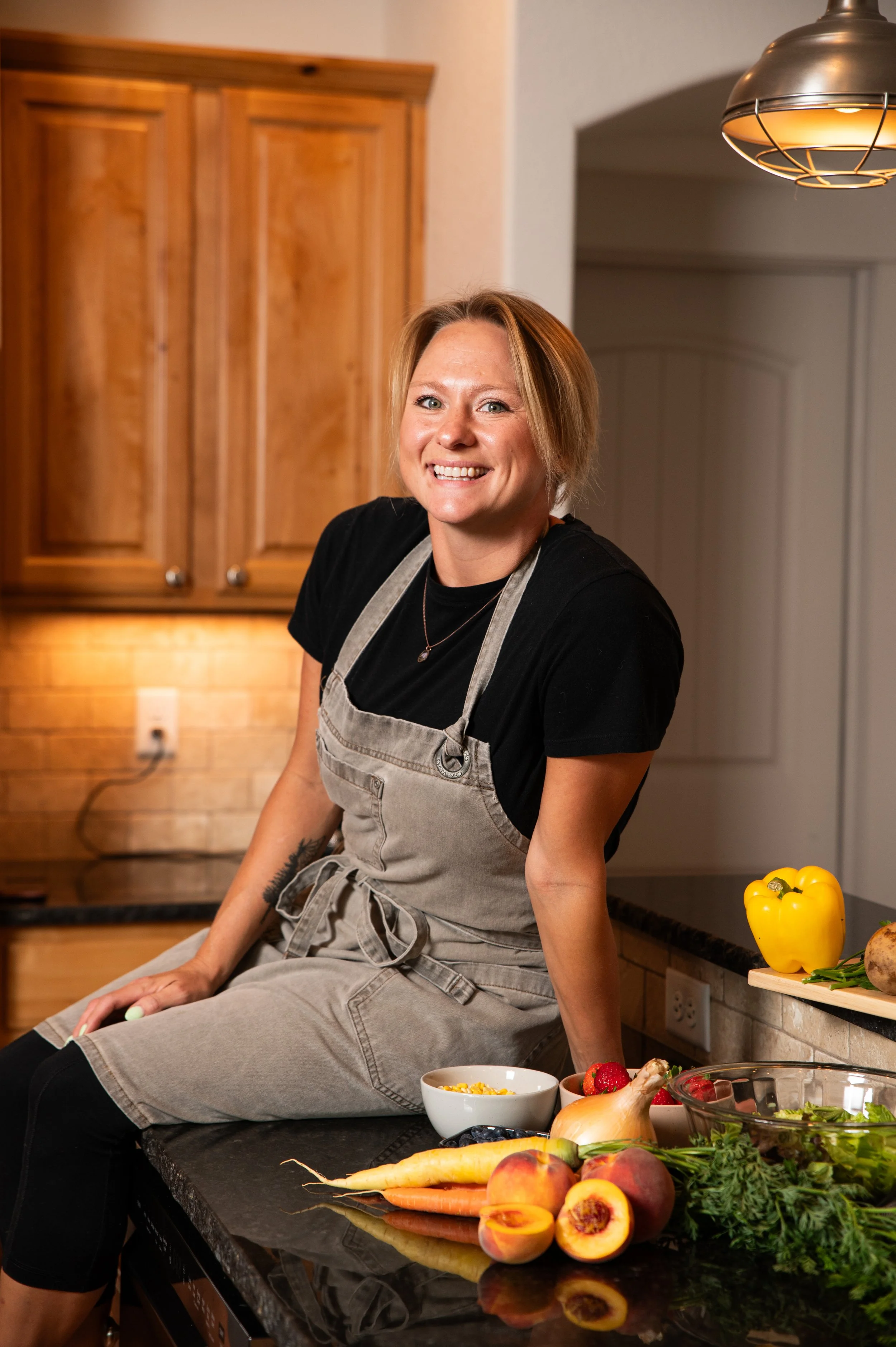 The owner, a female chef in a kitchen, is sitting on the counter, wearing an apron and smiling, with various fresh fruits and vegetables on the black countertop in front of her, including peaches, carrots, strawberries, and leafy greens.