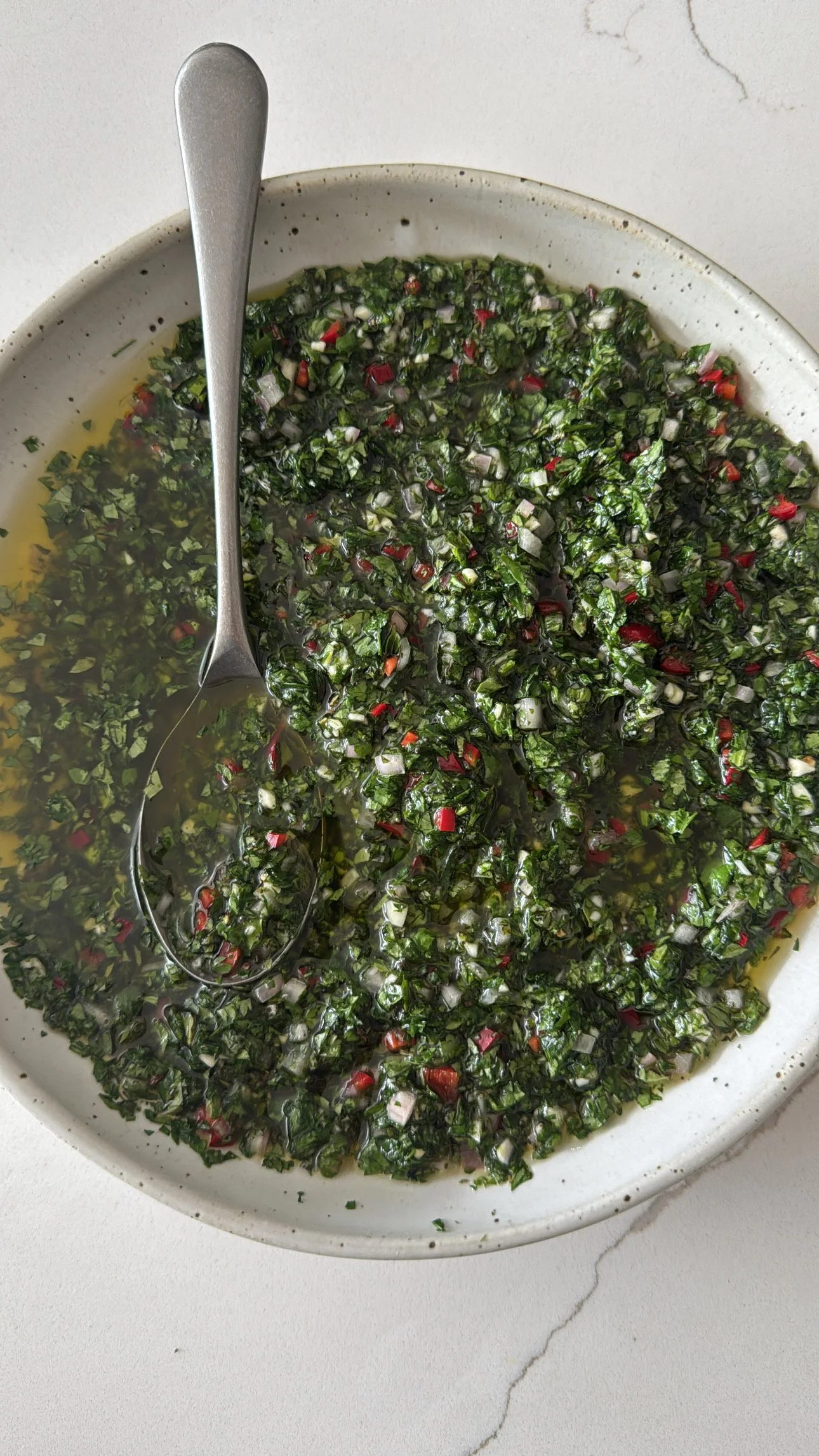 A white bowl containing chopped herbs, red chili, and oil, with a metal spoon in it, placed on a white surface with a marble pattern.