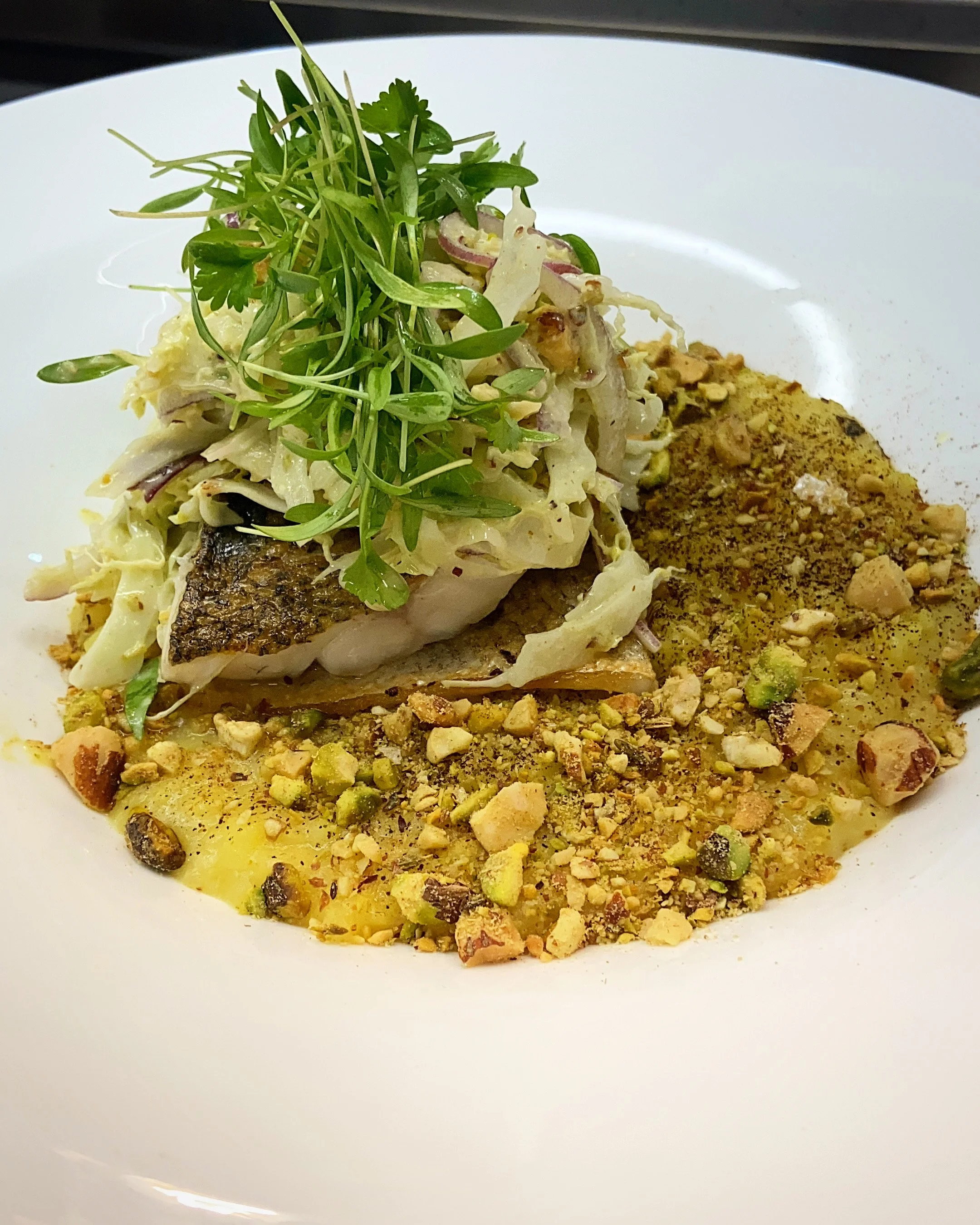 Close-up of a plated dish featuring a cooked fish fillet topped with microgreens and julienned vegetables, accompanied by a sprinkle of crushed nuts and a pile of ground spices on a white plate.