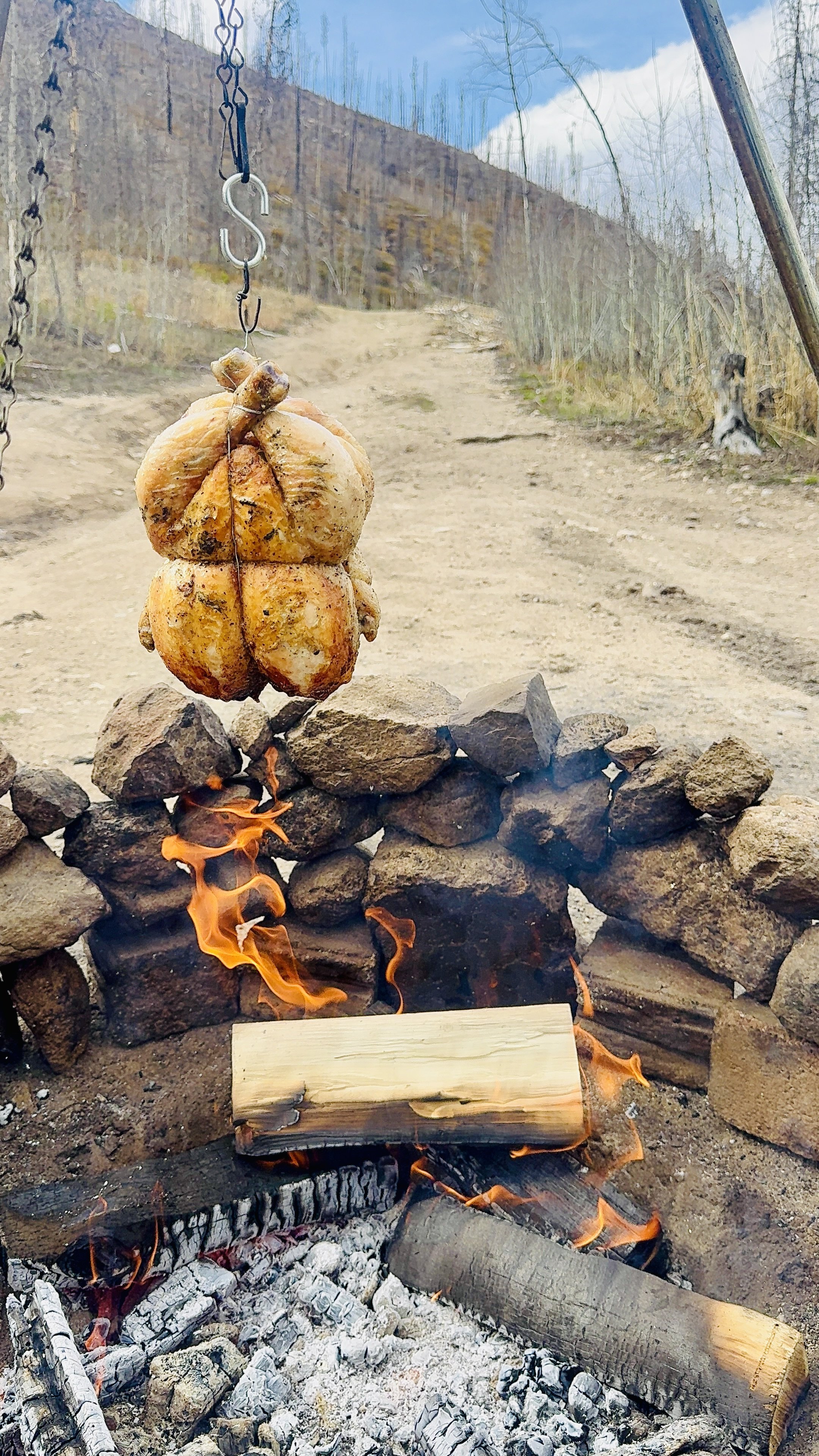A close-up of a campfire with stones around it, burning wood, and a chicken hanging on a chain above the fire, outdoors on a dirt path with trees and a clouded sky in the background.