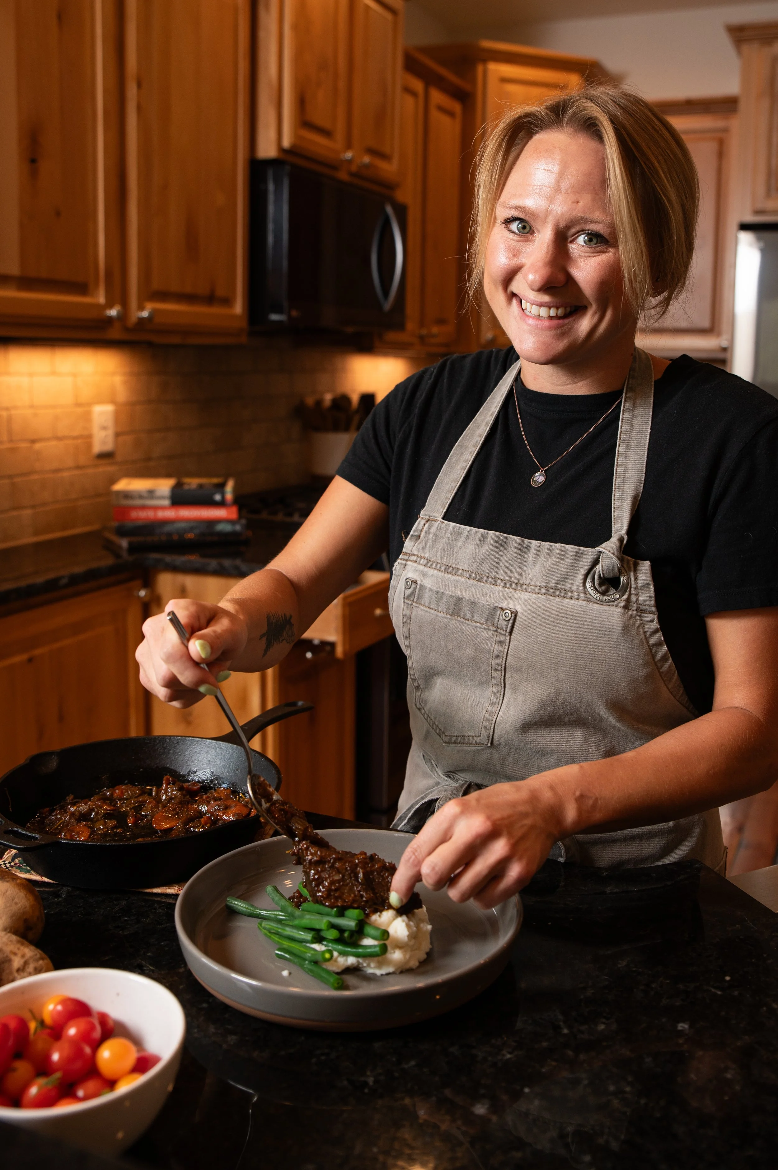 A woman with short blonde hair smiling while cooking in a kitchen, wearing a black shirt and beige apron. She is serving food onto a plate with mashed potatoes and green beans. There are cherry tomatoes in a bowl, books, and kitchen appliances visibl