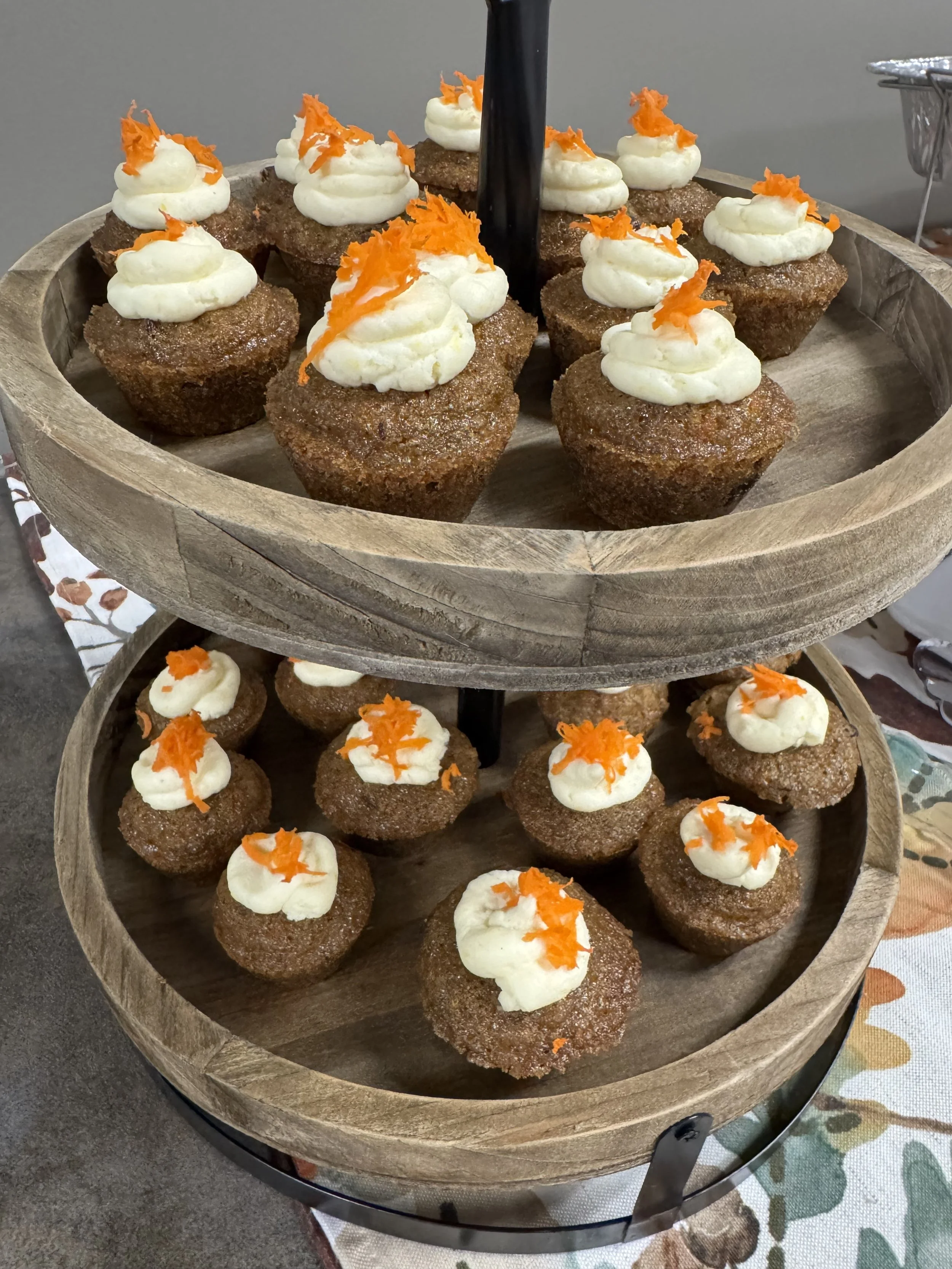 A two-tiered wooden stand with muffin cups topped with cream and orange shreds, displayed on a table with a decorative cloth.