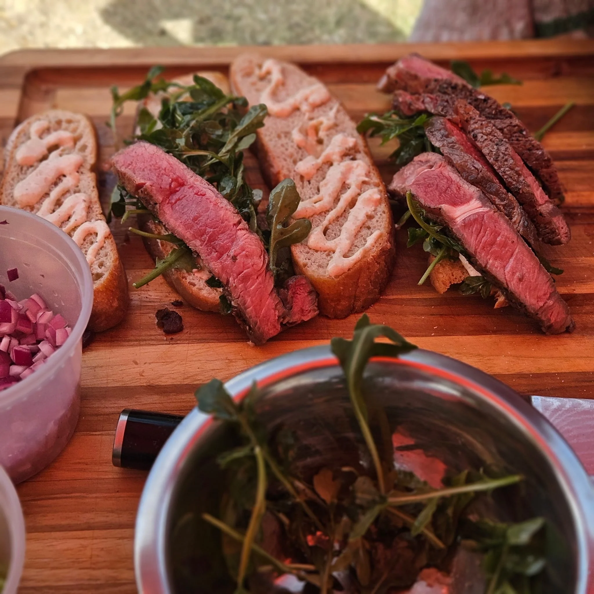 Close-up of slices of cooked steak arranged on a wooden cutting board with some arugula leaves. Part of a container of chopped onions and a cup are visible in the margins.