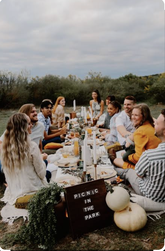 People having a picnic outdoors at a long table in a park setting with trees in the background, and a sign that reads "Picnic in the Park" with pumpkins and greenery decorations.