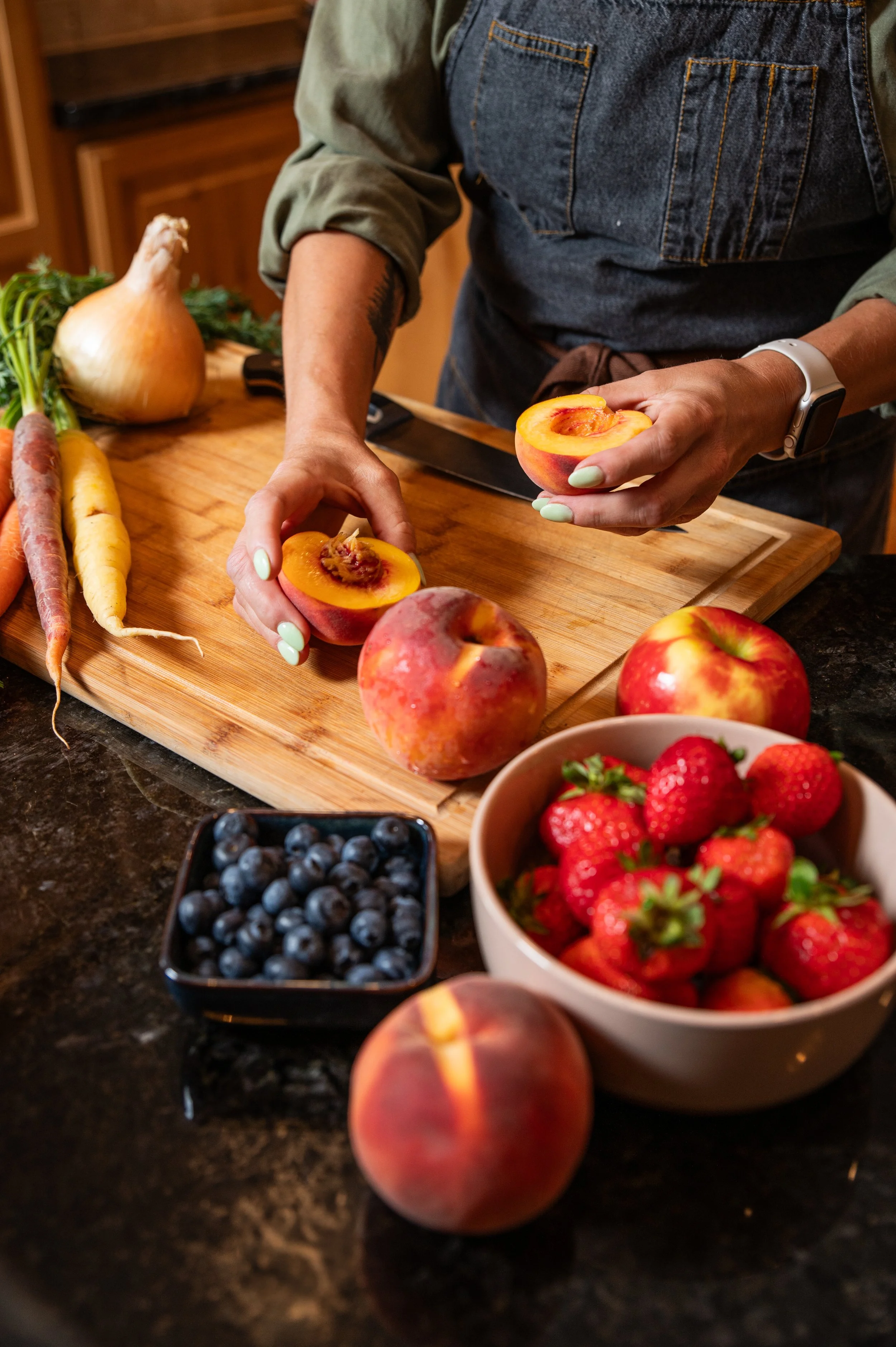 A person preparing fresh fruits and vegetables, including peaches, strawberries, blueberries, carrots, and an onion, on a wooden cutting board in a kitchen.