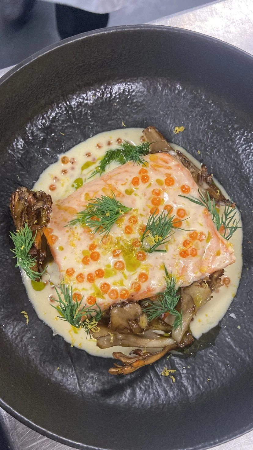 A gourmet dish featuring a poached egg on a bed of roasted vegetables in a creamy sauce, garnished with fresh herbs and orange and green roe, served in a black bowl.