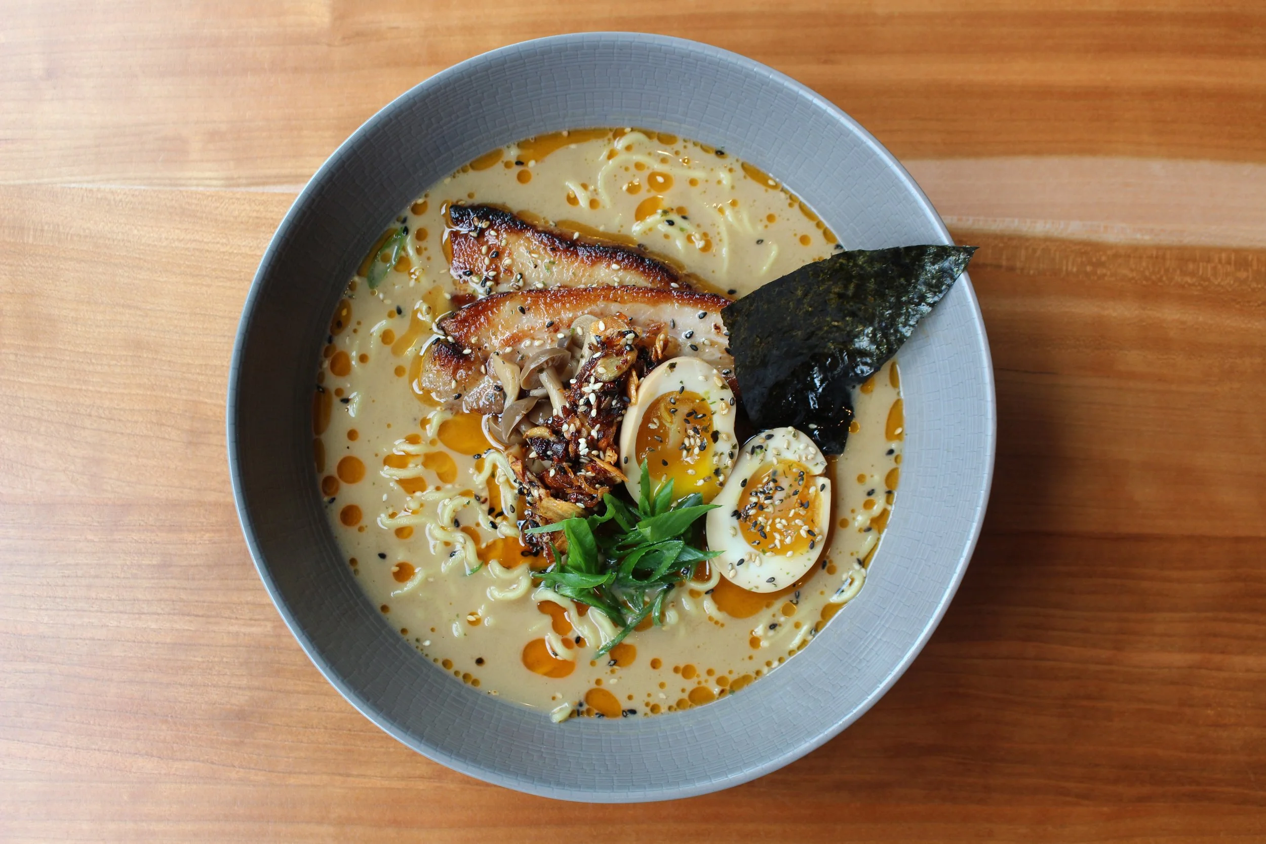 A bowl of ramen with slices of grilled pork, soft-boiled eggs, nori seaweed, green onions, mushrooms, sesame seeds, and chili oil on a wooden table.