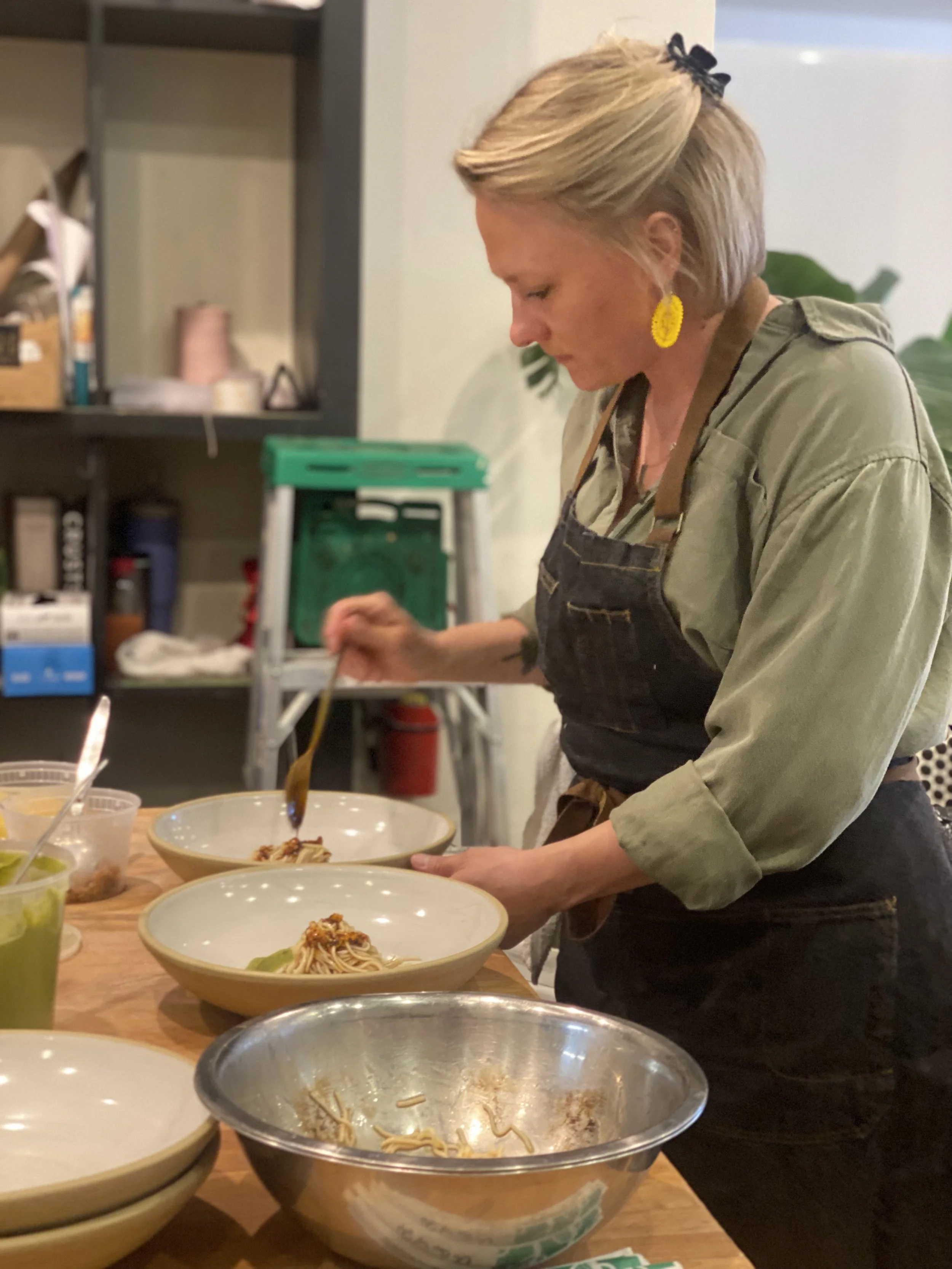 A woman with short blonde hair, wearing yellow earrings and a black bow in her hair, is preparing or serving food in a kitchen or dining area. She is focused on her task, standing behind a table with several bowls of food, including noodles and salad