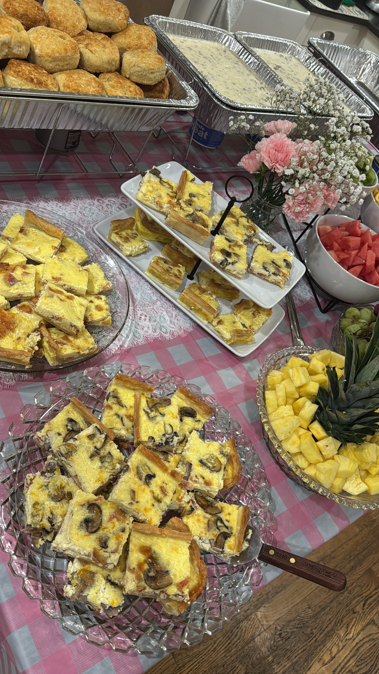 A buffet table with various dishes including chicken salad sandwiches, dish with creamy casserole, and fruit including watermelon, pineapple, and grapes, decorated with flowers.
