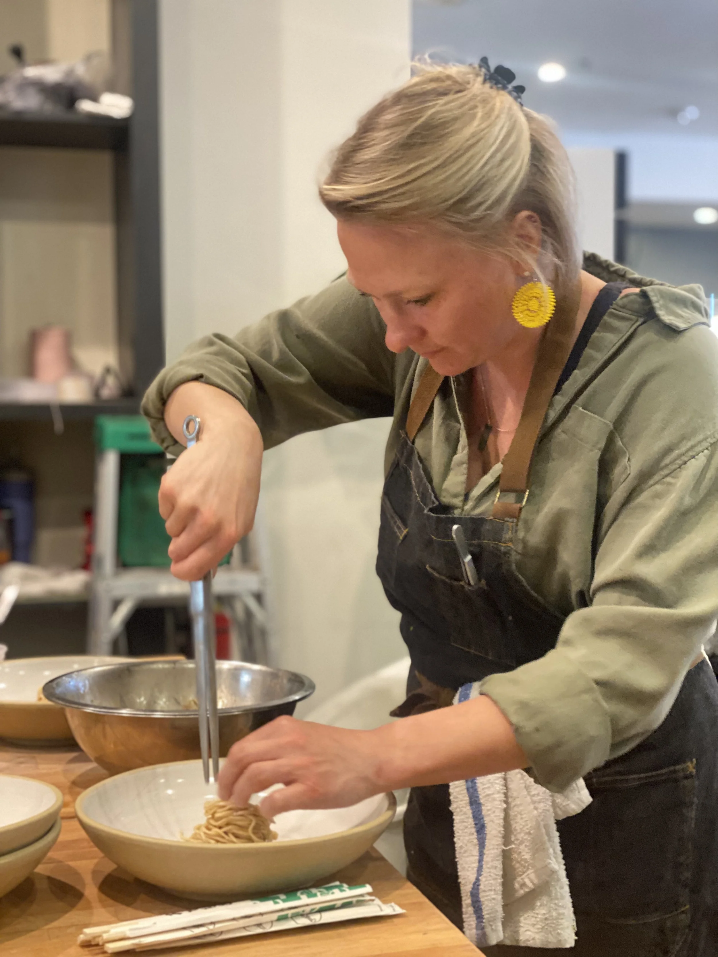 A person wearing a black apron and a green shirt is preparing to serve noodles into a ceramic bowl. The person has blonde hair tied back with a black hair accessory and yellow earrings. There are additional bowls on the table and kitchen shelves in t