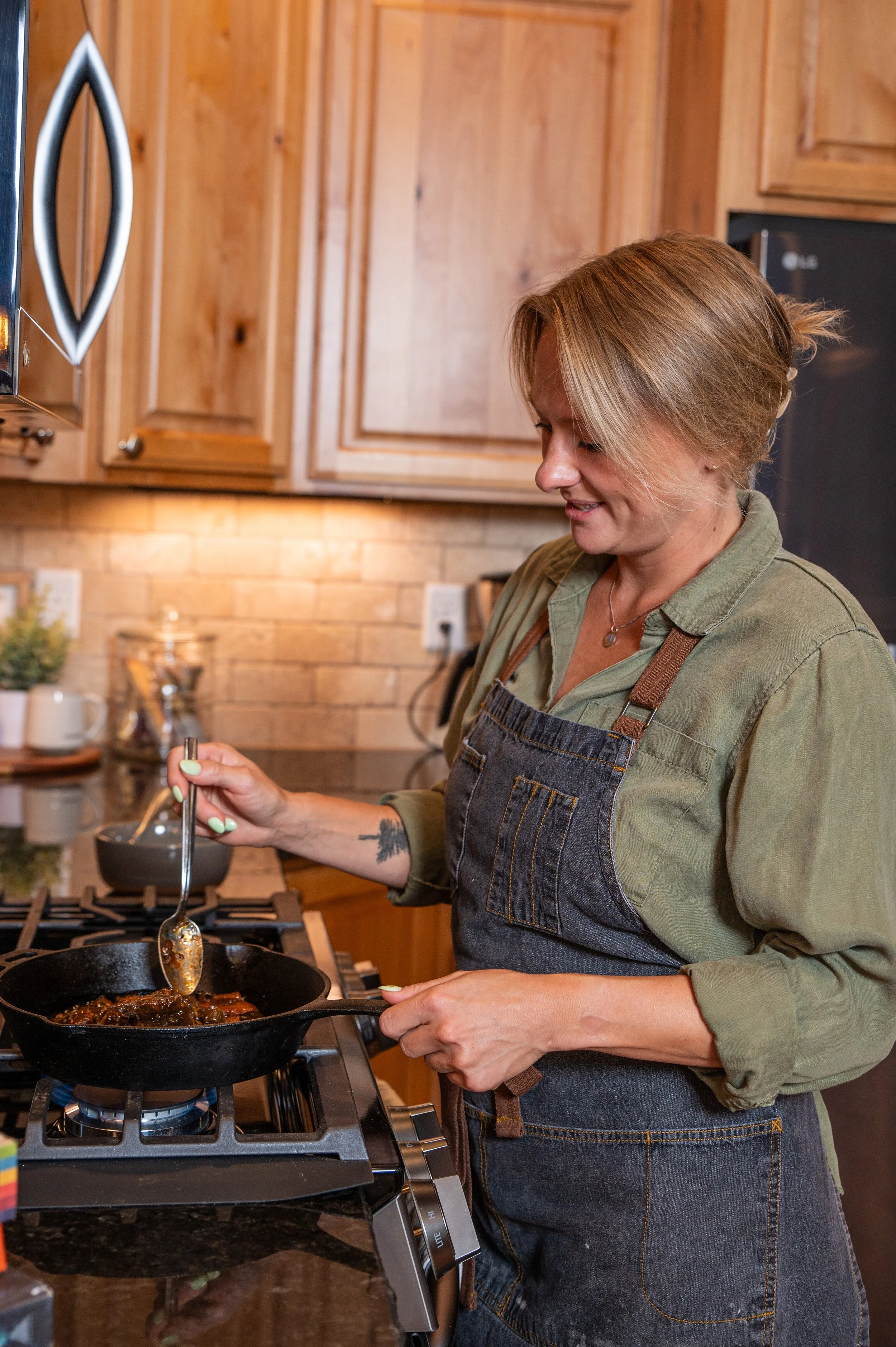 A woman with short, light brown hair cooking on a stovetop in a kitchen with wood cabinets and a brick backsplash. She is wearing a green shirt and a denim apron, stirring food in a black skillet.