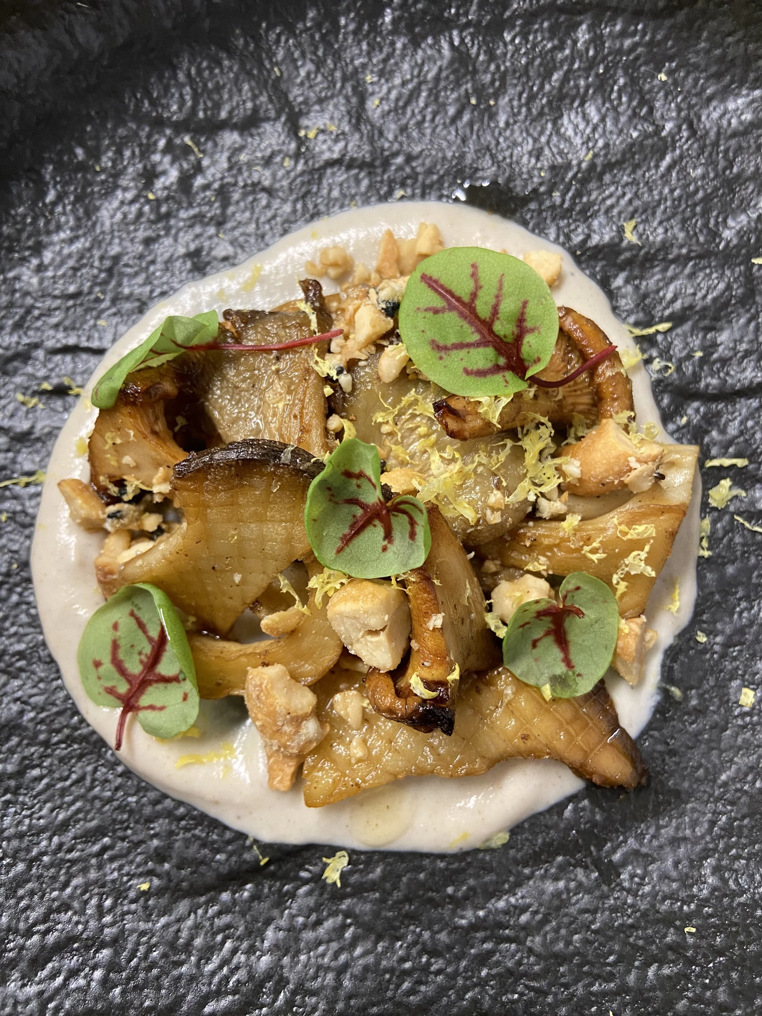 Close-up of a gourmet dish featuring roasted or grilled vegetables, microgreens, and crumbled nuts on a creamy white sauce or base, on a dark textured surface.