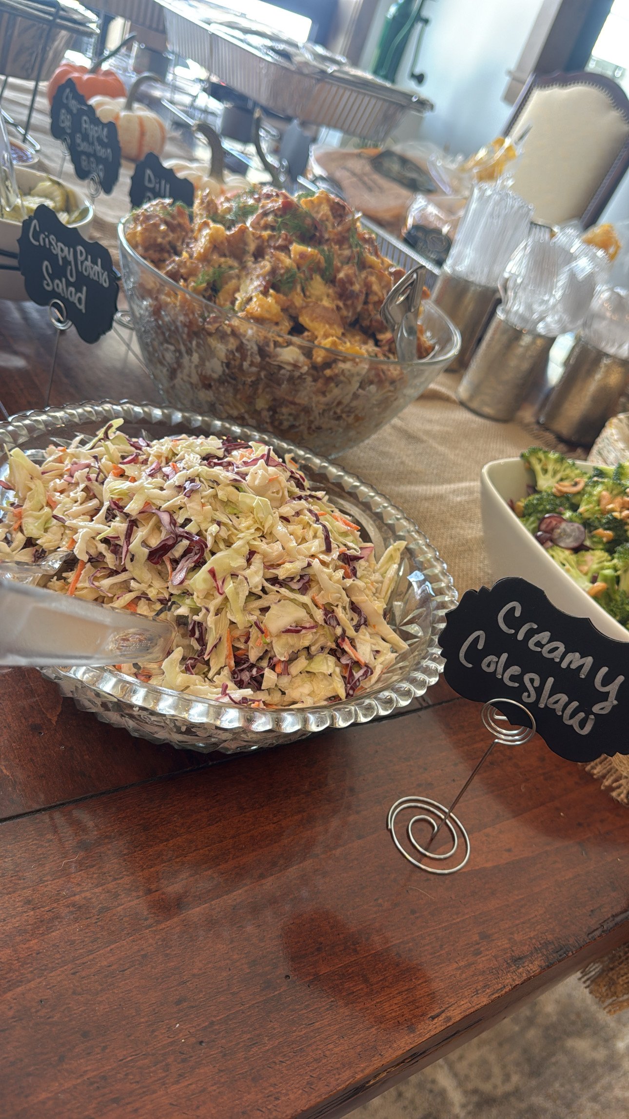 A buffet table with various salads and dishes, including coleslaw, potato salad, and a dish labeled 'Creamy Cleslaw'.