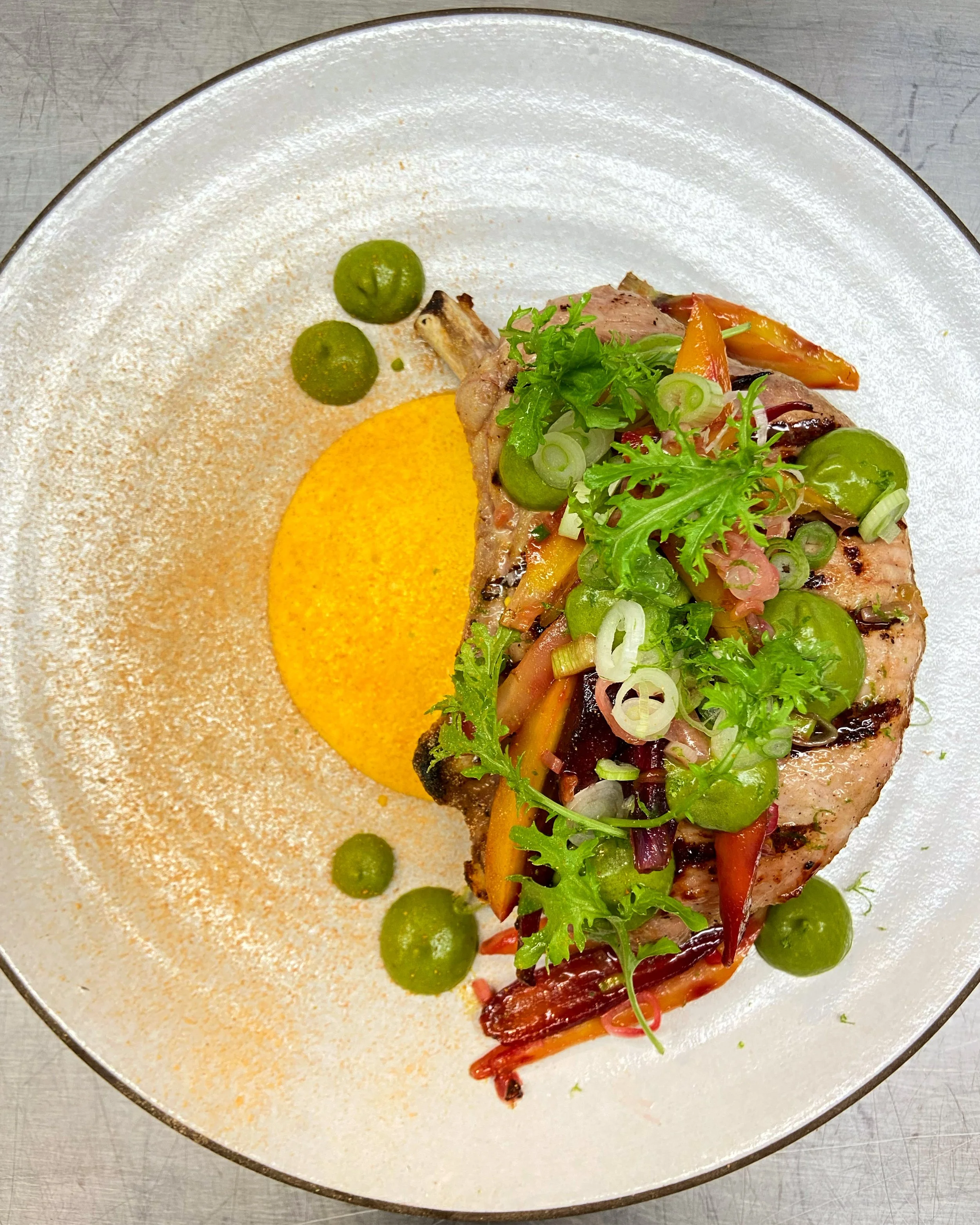 Close-up of a plated dish featuring grilled meat topped with fresh green herbs, sliced scallions, and colorful roasted bell peppers, served with a yellow sauce and green pea garnishes on a white oval plate.