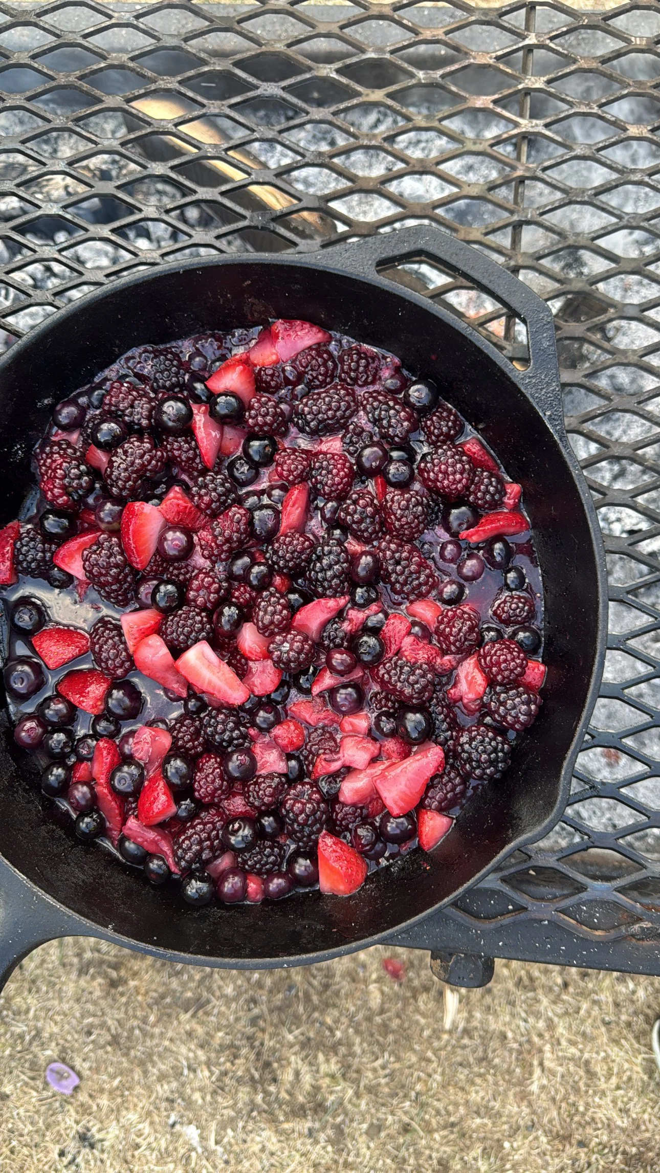 A black skillet containing a mixture of blackberries, sliced strawberries, and blueberries, with a dark liquid, possibly syrup or juice, in the skillet. The skillet is placed on a metal outdoor table with a diamond pattern, and the ground beneath is 