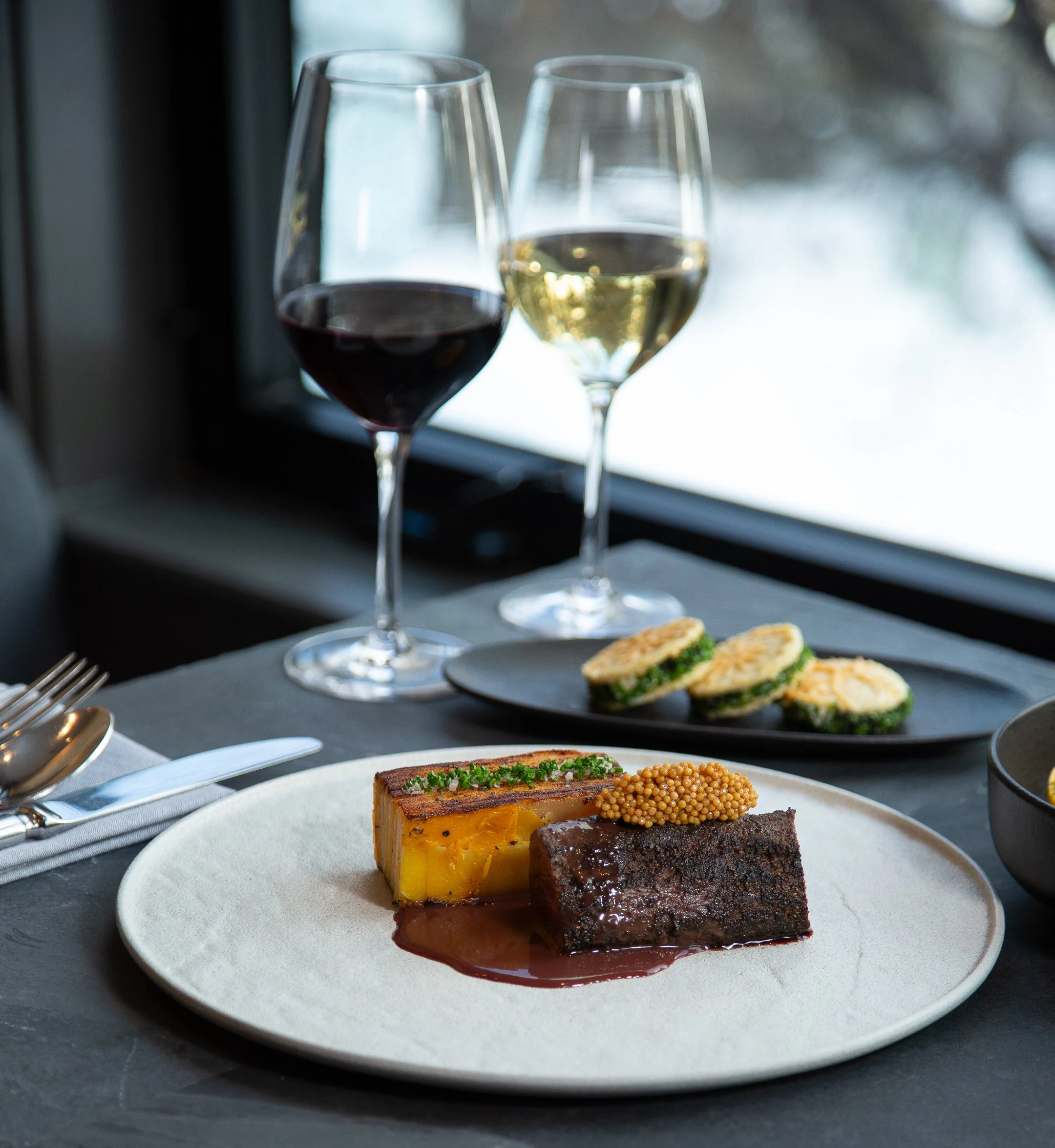 Close-up of a white plate with three slices of layered dessert topped with sauce and garnished with herbs, on a black table. In the background, there are two wine glasses, one with red wine and one with white wine, on a table set for dining.