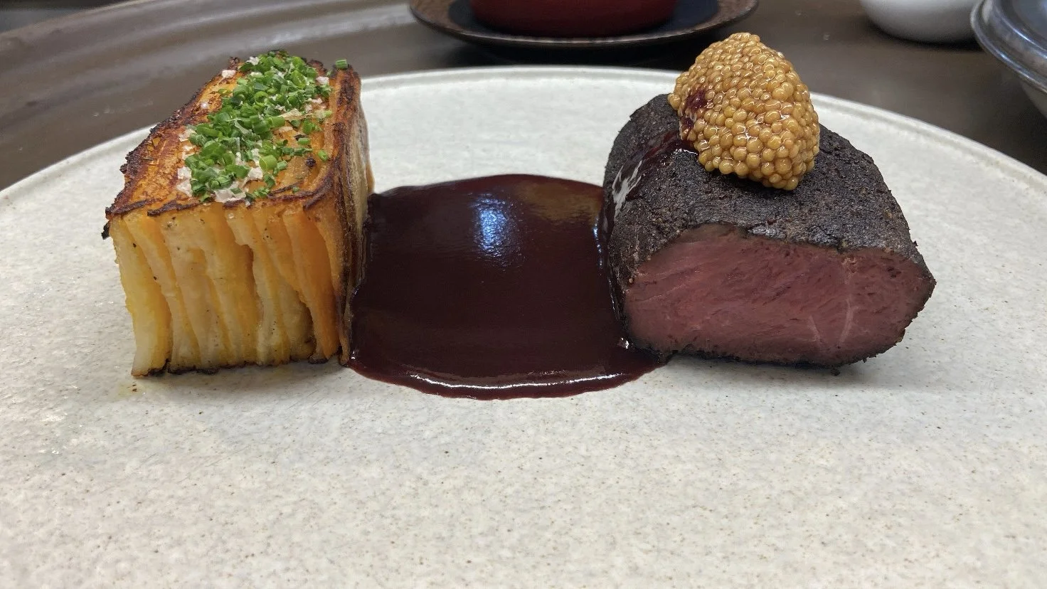 A plate with a piece of beef tenderloin topped with mustard seeds and a side of potato gratin garnished with chopped chives, with dark sauce served on the plate.