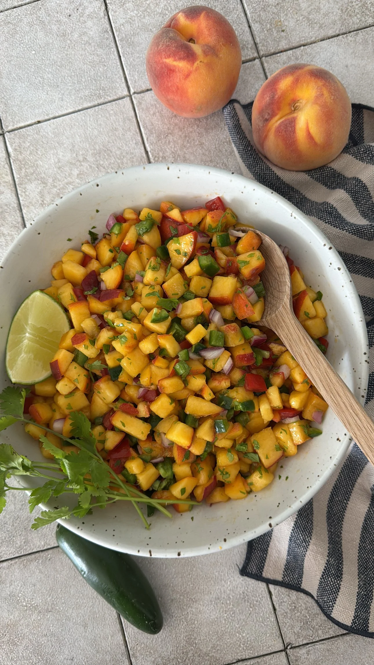 A white bowl filled with diced yellow mango, red bell pepper, chopped red onion, and chopped green herbs, garnished with cilantro and a lime wedge. There are two peaches and a cucumber on the tiled surface nearby, with a striped cloth and a wooden sp