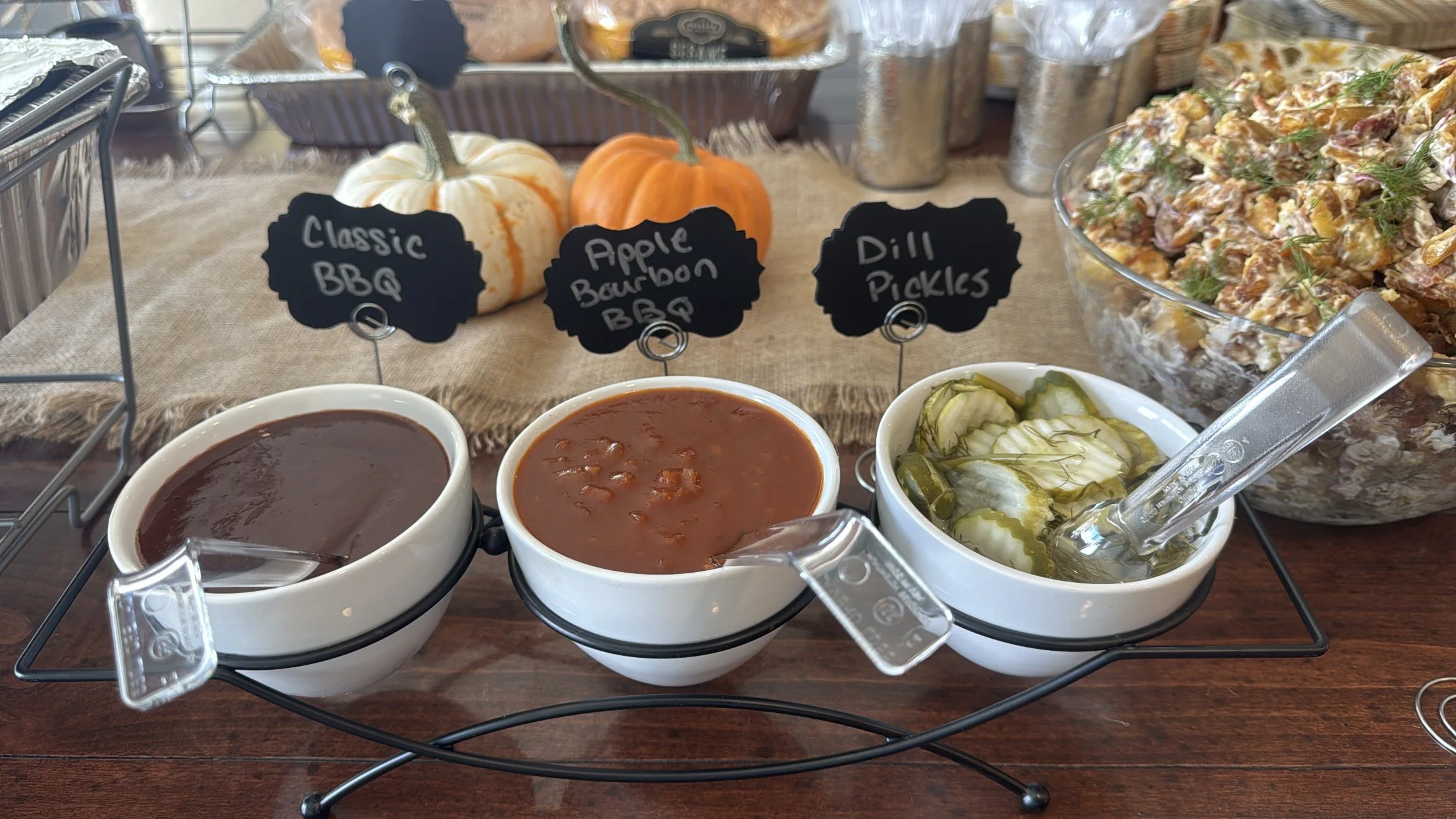 A buffet table with three bowls of different sauces or soups, two small pumpkins, and a large bowl of a creamy salad with herbs. There are also small pickles and signs indicating the items: 'Classic BBQ,' 'Apple Bourbon BBQ,' and 'Dill Pickles.'