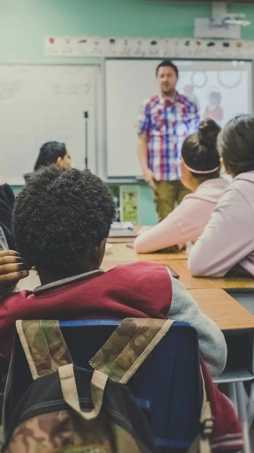 Students sitting at desks in a classroom, watching a teacher who is standing in front of a whiteboard and a digital display.