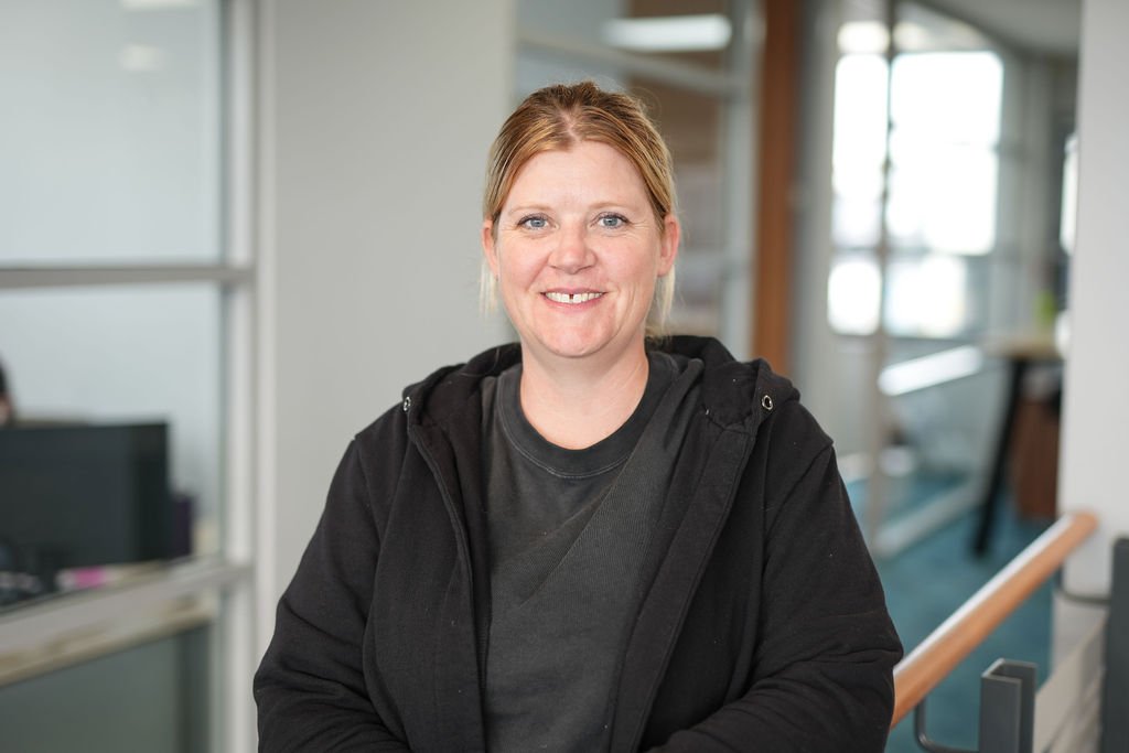 A woman with blonde hair wearing a black hoodie standing indoors with a background of windows and office furniture.