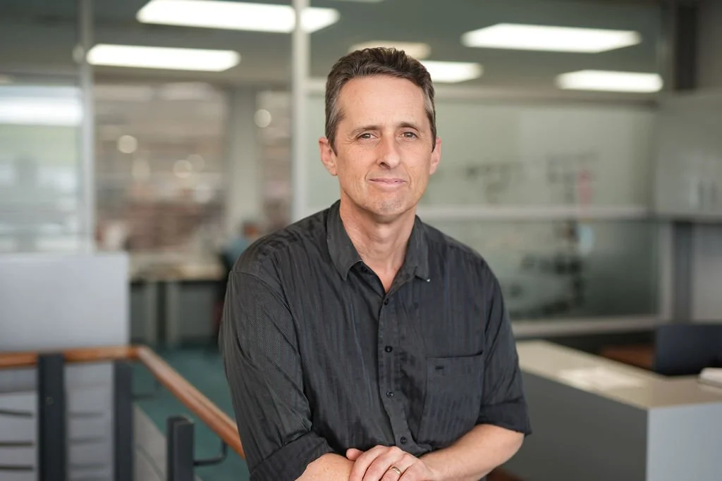 A man with short dark hair wearing a black shirt standing with arms crossed in an office environment.